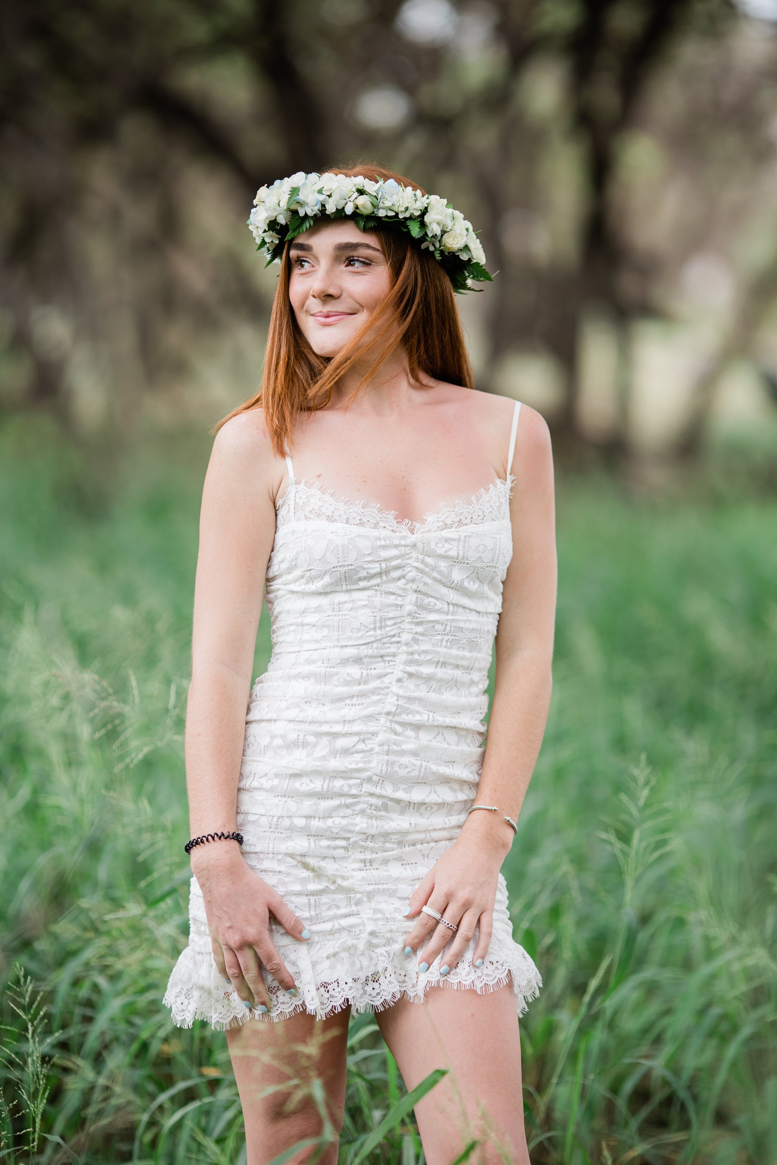 A young woman with long red hair wearing a white lace dress and a floral crown of white flowers standing outdoors in a green setting with trees.