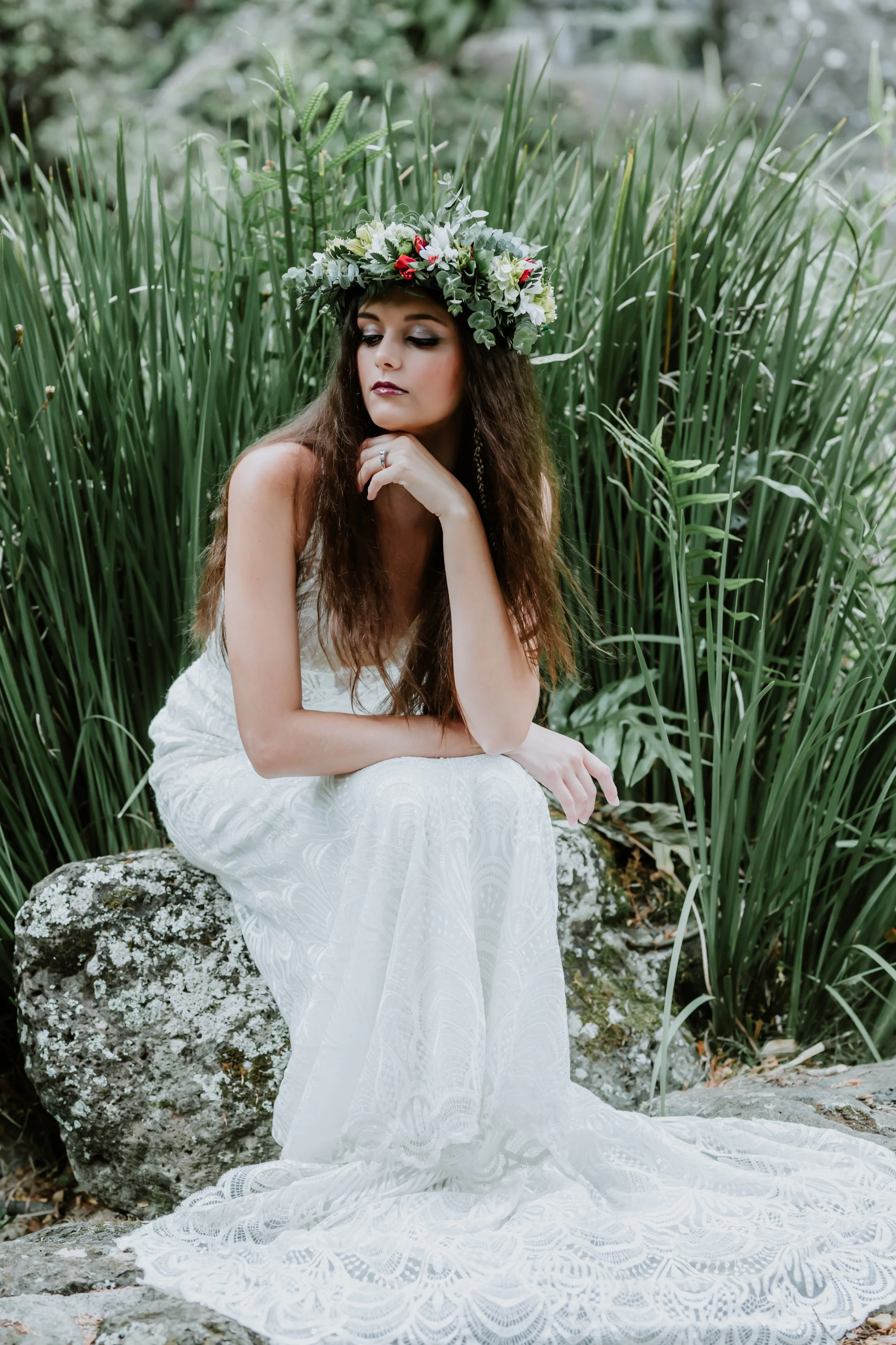 A woman sitting on a rock outdoors, wearing a white lace dress and a floral crown, surrounded by tall green grass and rocks.