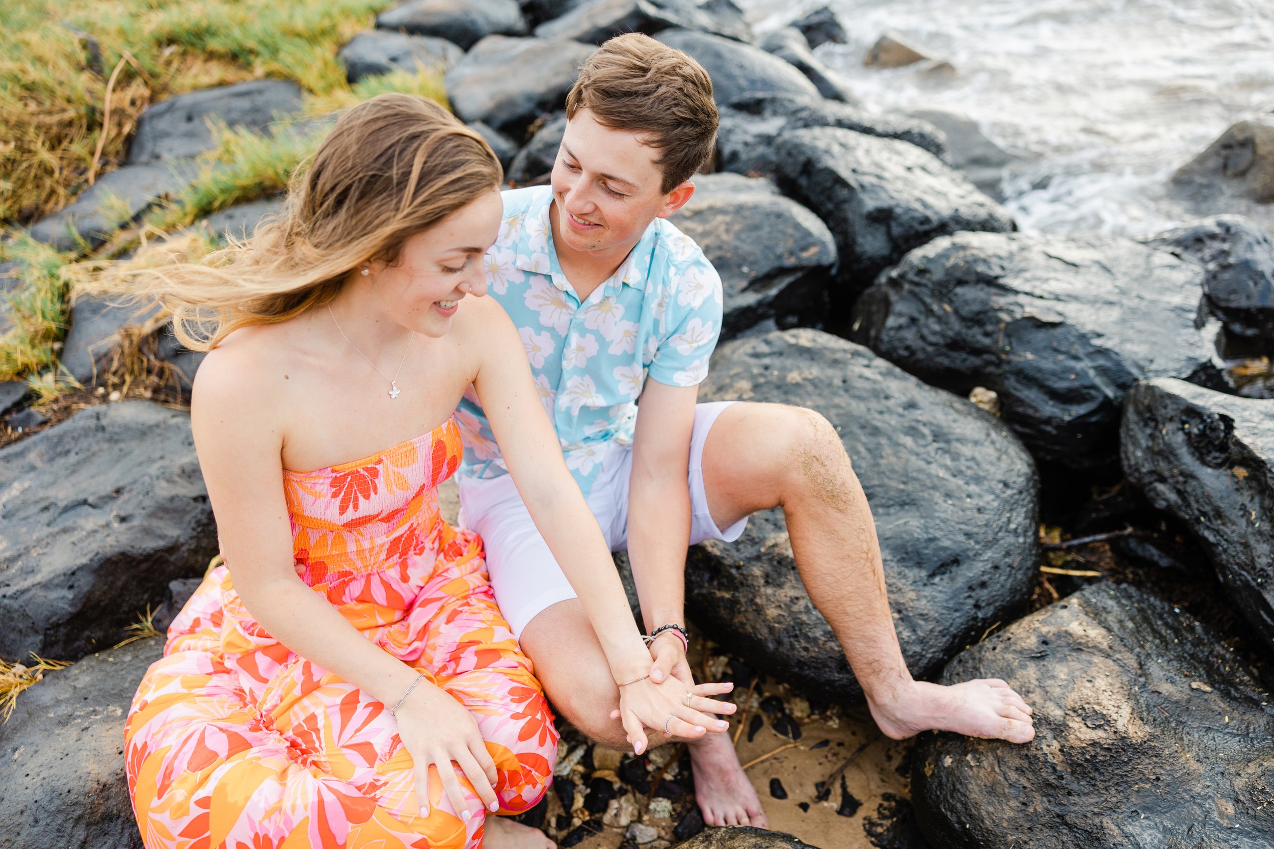 A young man and woman sitting on rocks by the water, smiling and enjoying each other's company on a beach.