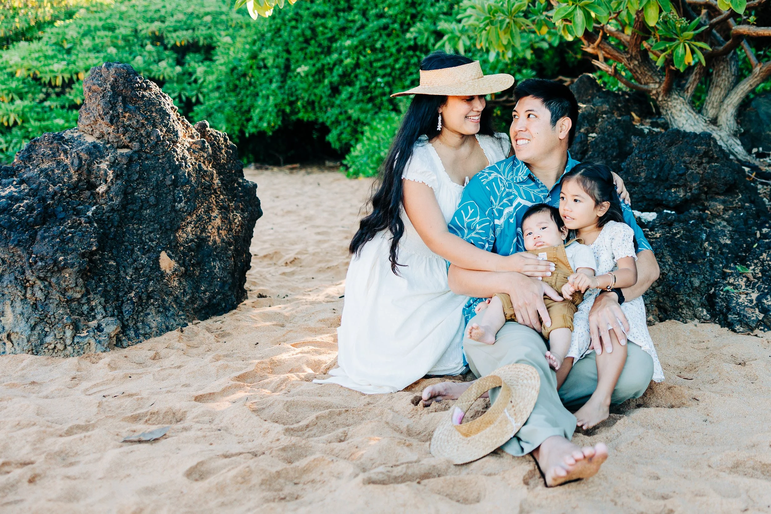 Family of four sitting on the sandy beach, with rocks and green foliage in the background. The mother wears a white dress and straw hat, the father a blue shirt, and the children, a little girl in a white dress and a baby in brown overalls, are sitting on the father's lap.