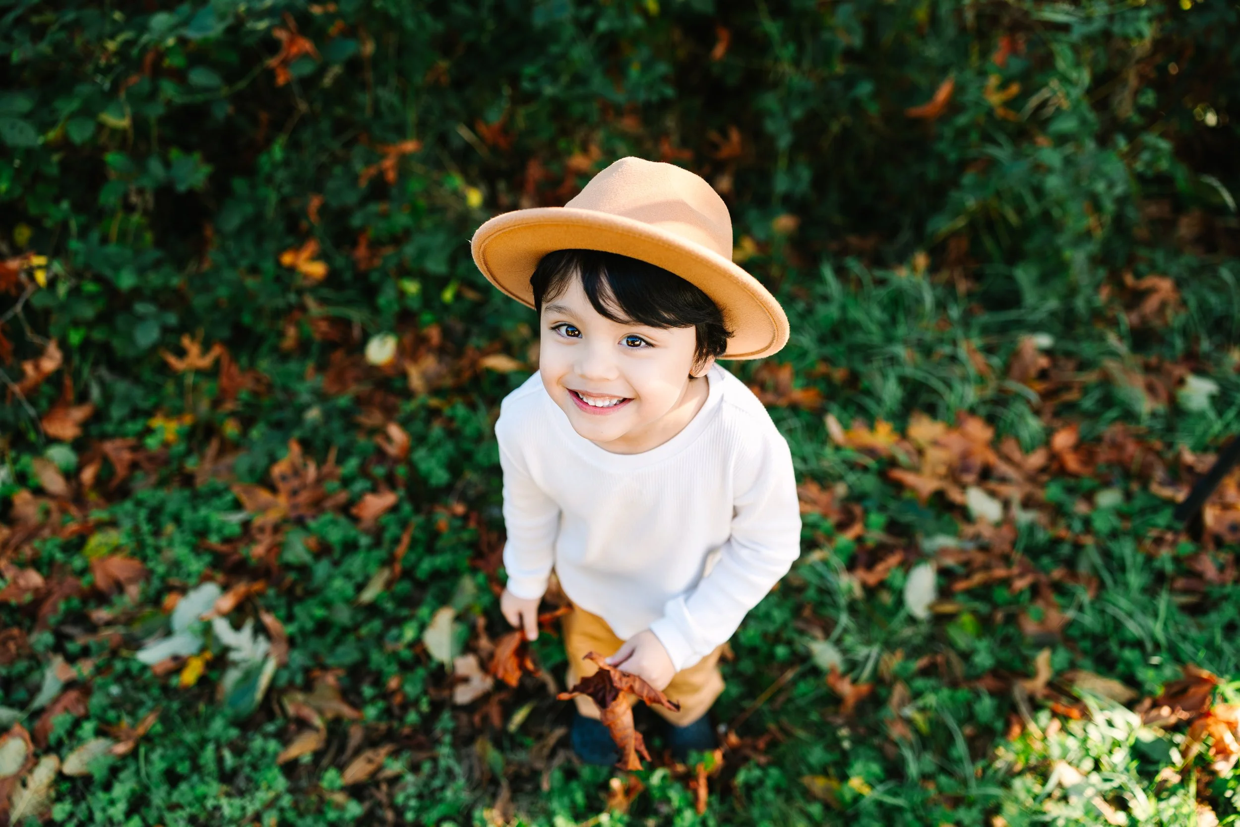 A young boy with a beige hat, white shirt, and tan pants smiling while standing outdoors on fallen leaves and green foliage.