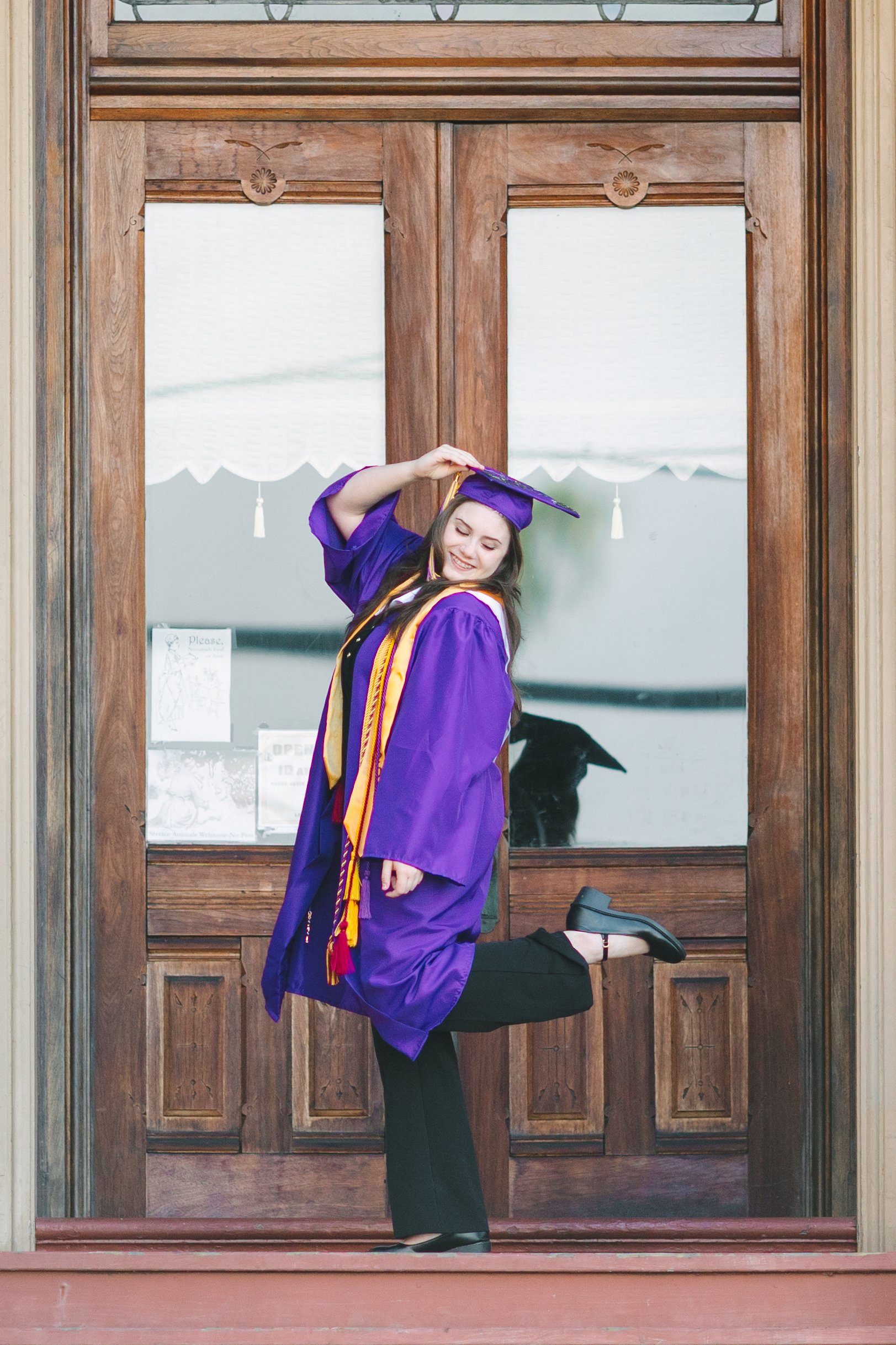 A young woman in a purple graduation gown and cap, with a gold stole, smiling and striking a playful pose in front of a wooden door.