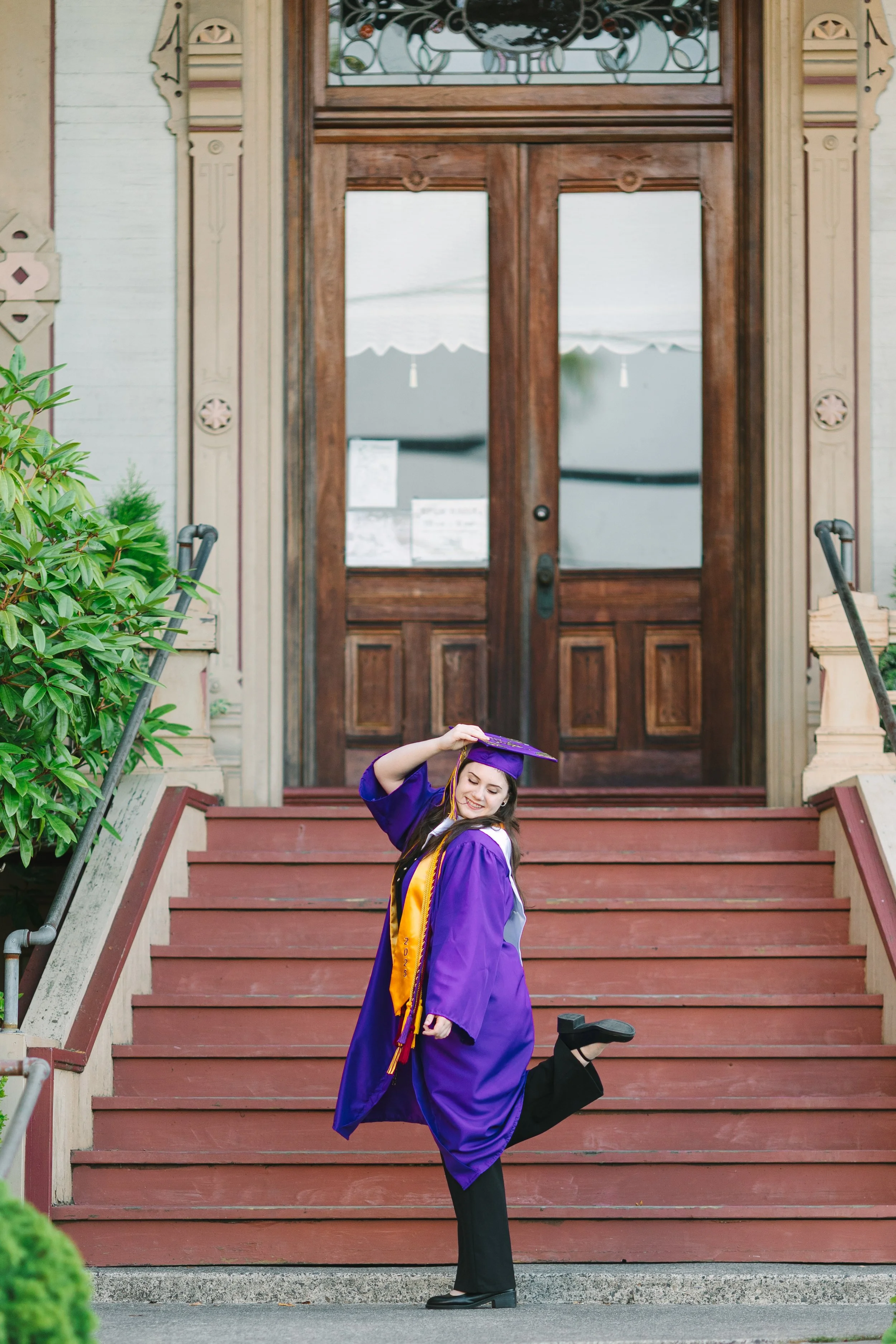 A young woman in a purple graduation gown and cap with yellow and purple cords, smiling and posing on a red wooden staircase in front of a large wooden door with glass panels. She is lifting her right leg and holding her cap with her right hand, stan