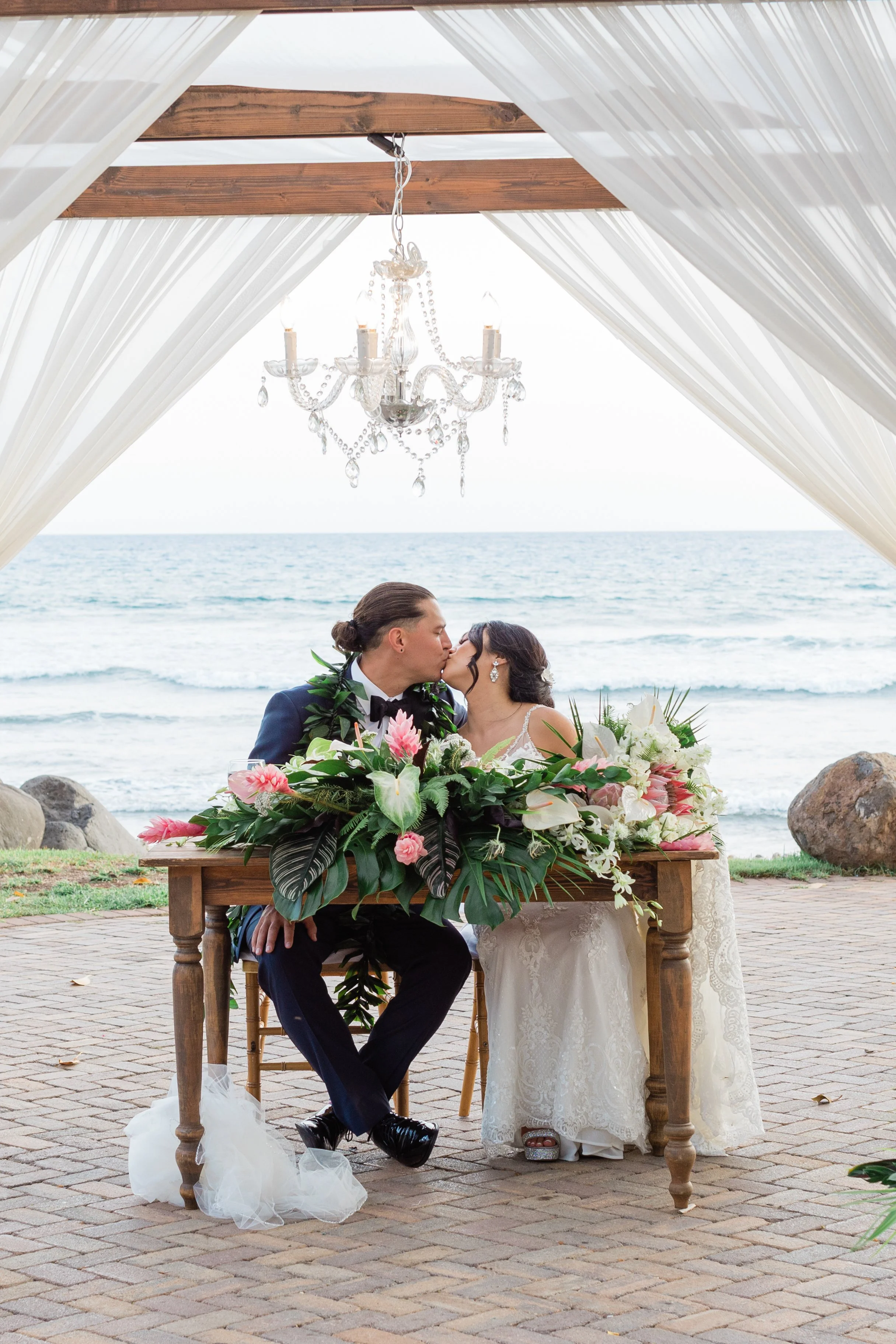 A bride and groom are sitting and kissing at their wedding table on the beach, surrounded by floral arrangements and under a canopy with draped white fabric and a chandelier, with ocean waves in the background.