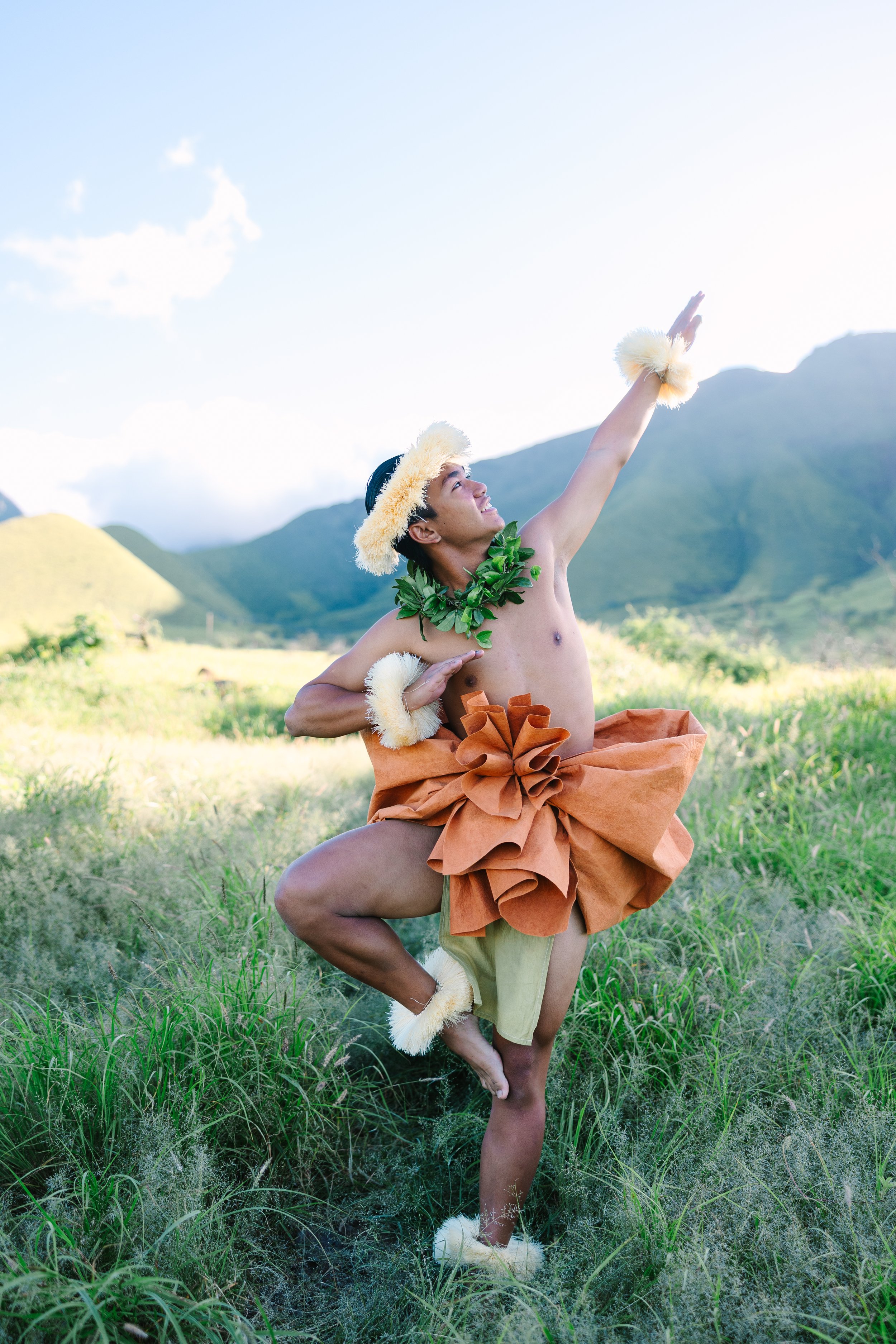 Person dressed in traditional Polynesian attire balancing on one leg in green field with mountains in background