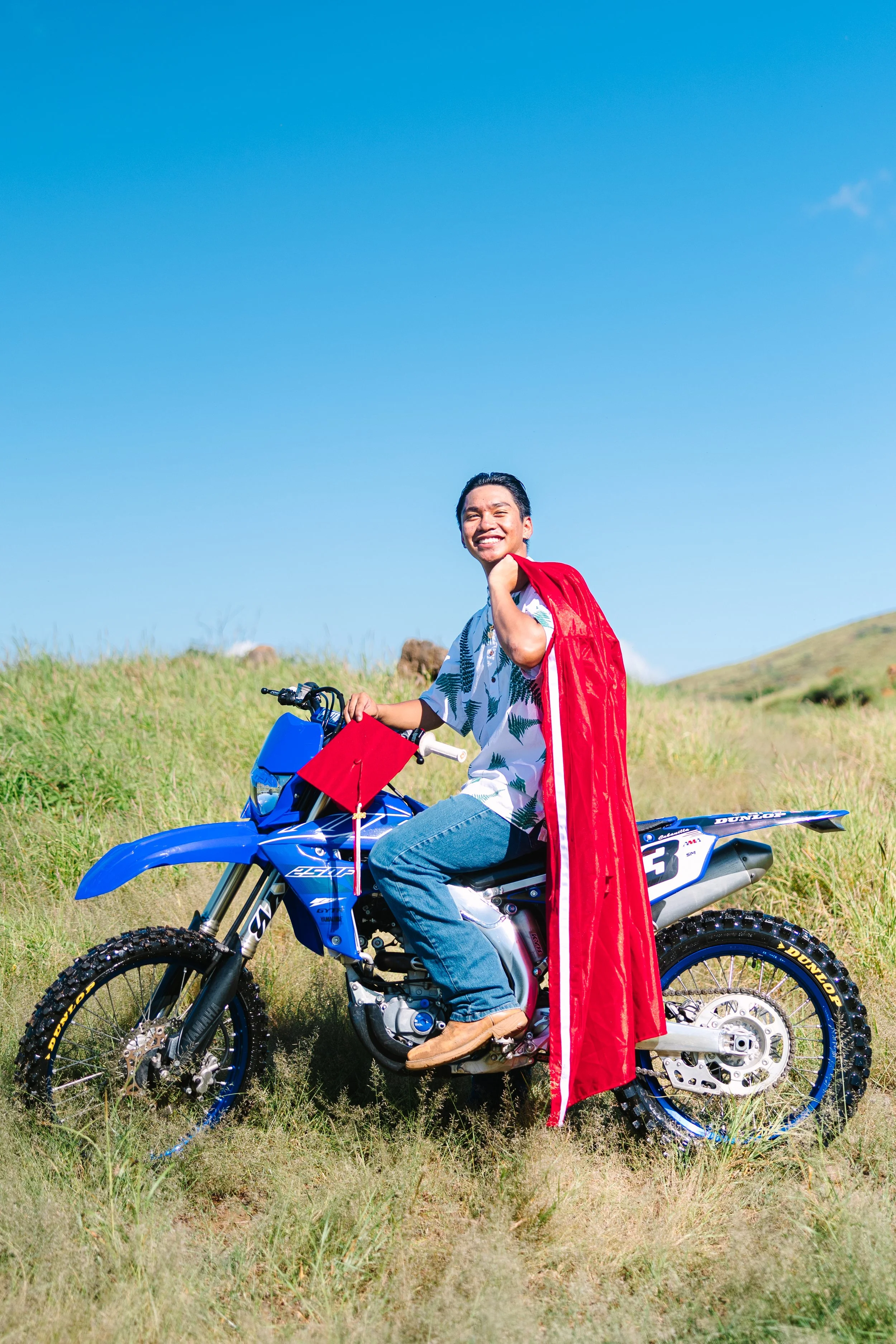 A person smiling while sitting on a blue dirt bike in an open grassy field under a clear blue sky, wearing a red cape over a Hawaiian shirt and jeans.