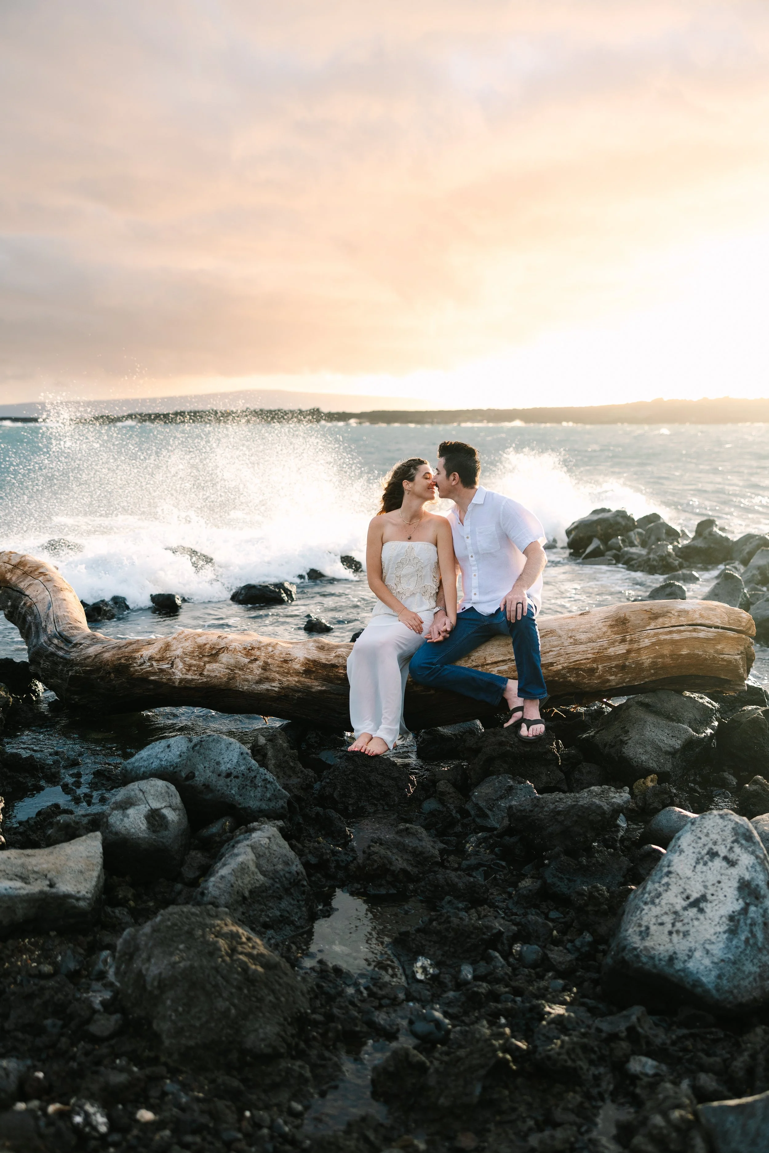 A Maui couple sits on a large driftwood log near the rocky shoreline with ocean waves crashing behind them. This engagement session a one to remember. The scene is illuminated by the setting sun, creating a warm, romantic atmosphere.