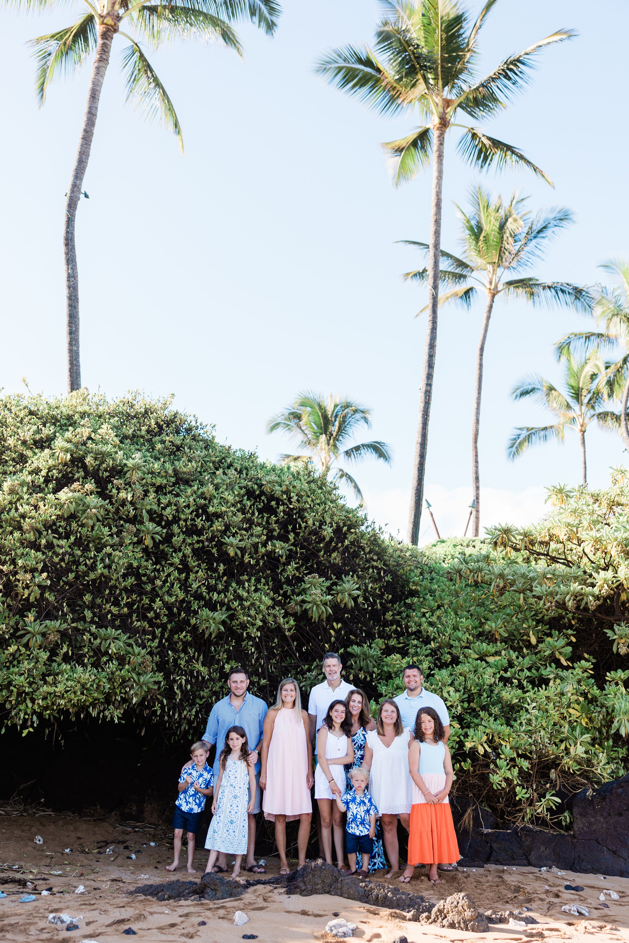 A large family posing on the beach in front of lush green foliage and tall palm trees, with the ocean and bright sky in the background. Poolenalena Beach in Maui is so beautiful and ideal for early morning photo shoot. 