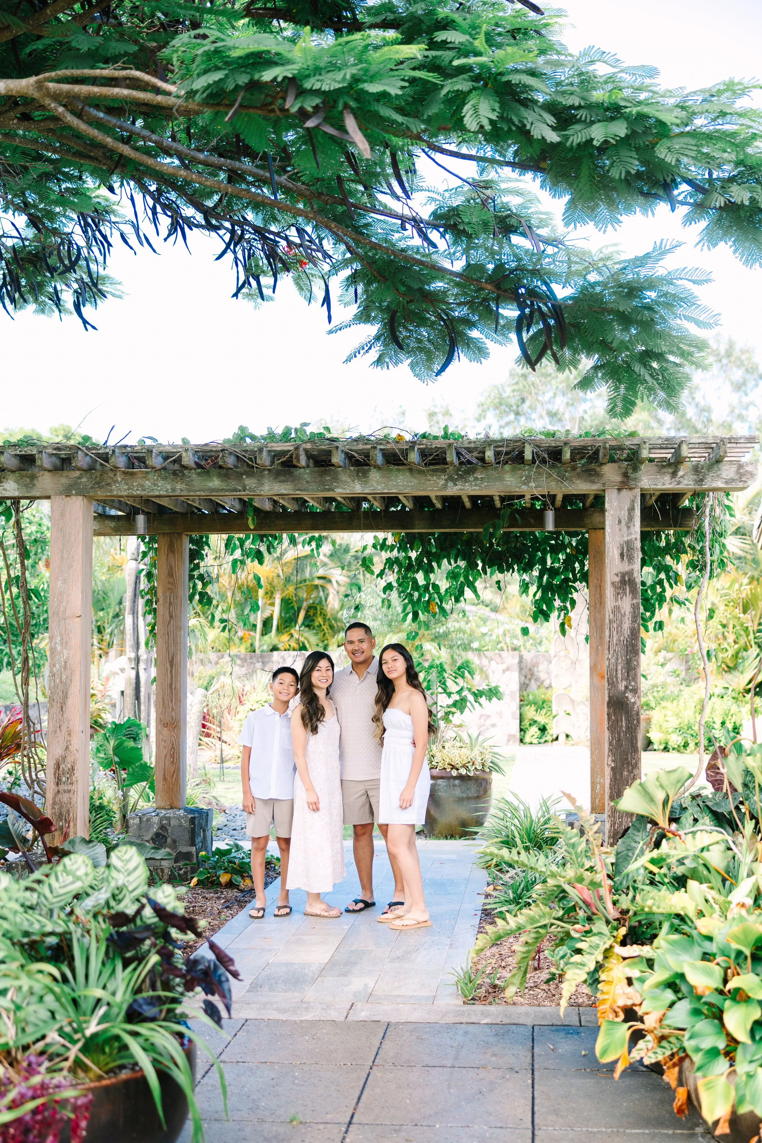 Family of four posing under a wooden pergola in a lush garden with green plants and trees. A wonderful Maui photo session. 