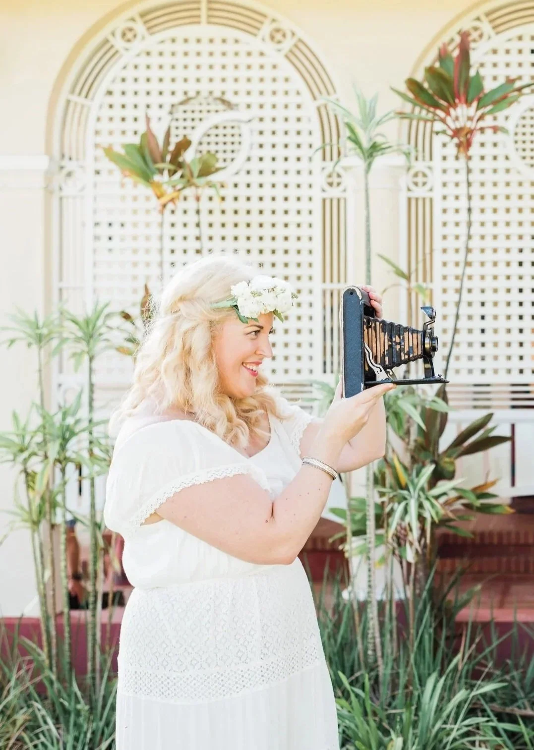 A woman with blonde hair, wearing a white dress and a flower crown, is smiling and taking a picture with an old-fashioned camera indoors, behind her is a decorative lattice wall with tropical plants.