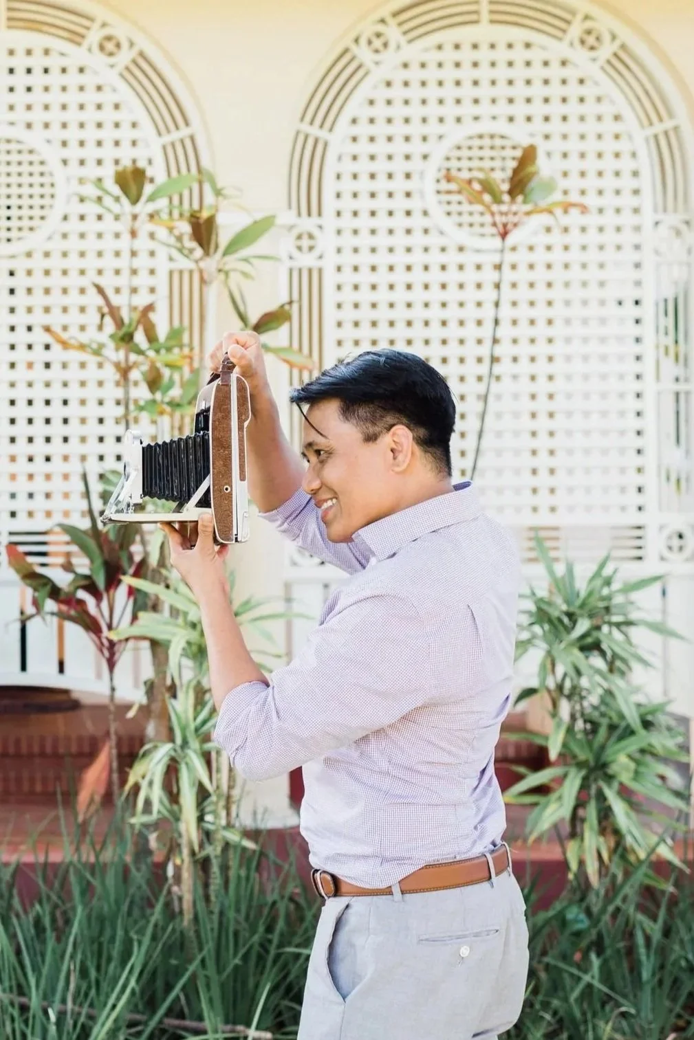 Smiling man in light gray pants and a light purple button-up shirt taking a photograph with a vintage camera outdoors, with green plants and a decorative white lattice background.