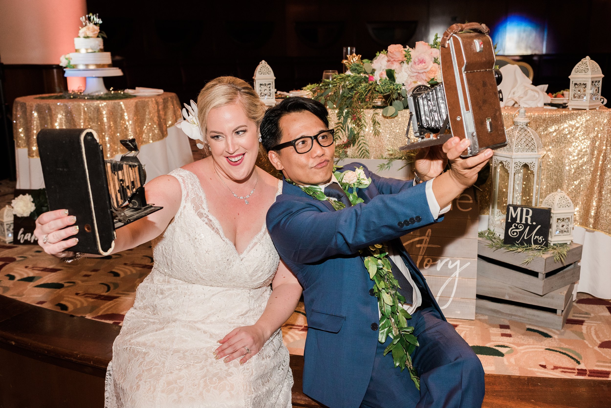 A bride and groom taking a selfie at their wedding reception. The bride is wearing a white lace wedding dress, smiling. The groom is in a blue suit with glasses and a green lei, holding a vintage camera, appearing focused on the selfie.