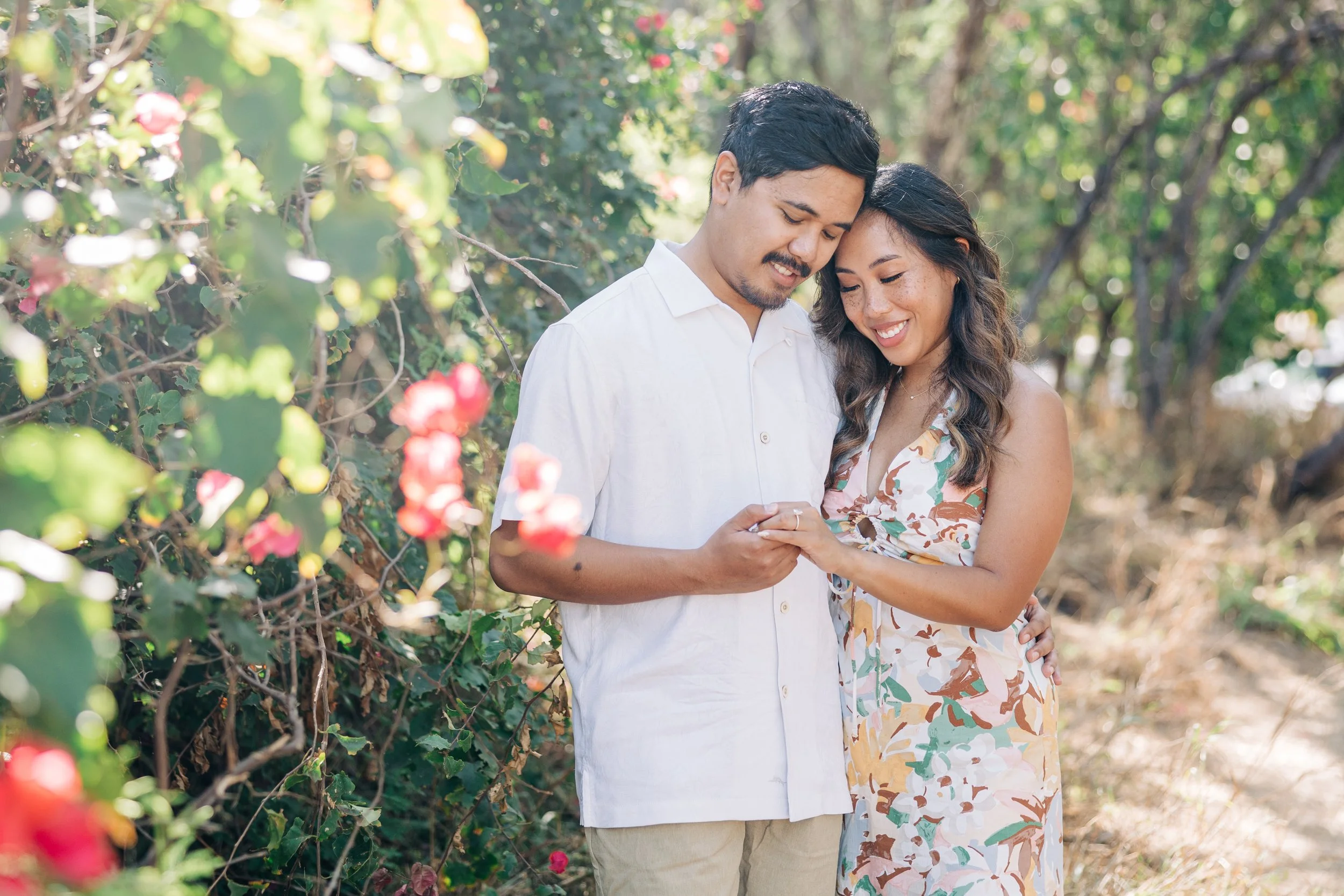 A Maui Engagement Photo Shoot. A smiling couple standing close together outdoors among greenery and pink flowers, with the man wearing a white shirt and the woman in a colorful floral dress.