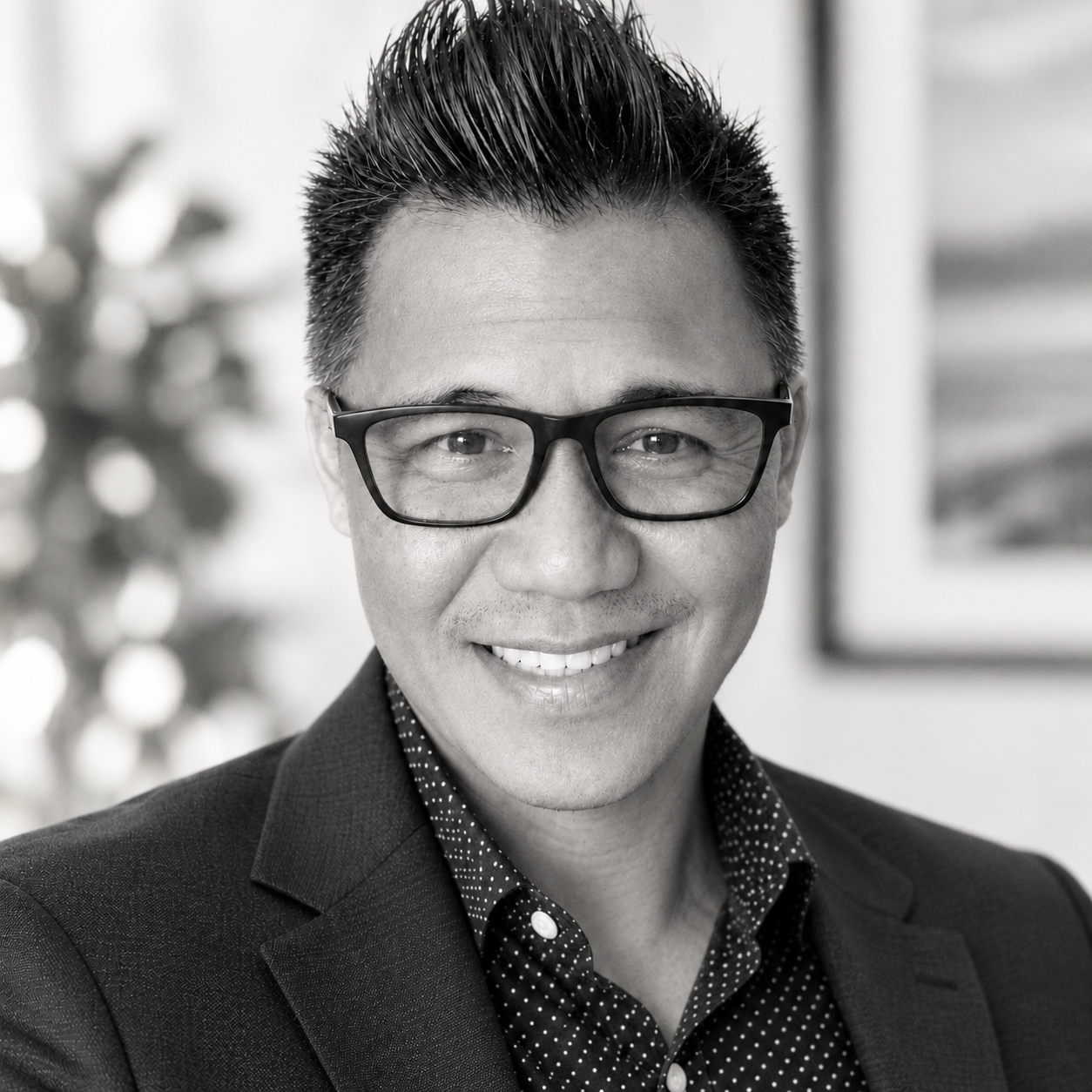 Meet your Maui Photographer, Rolland. Black and white portrait of a smiling man wearing glasses, a patterned shirt, and a blazer, with a blurred background.