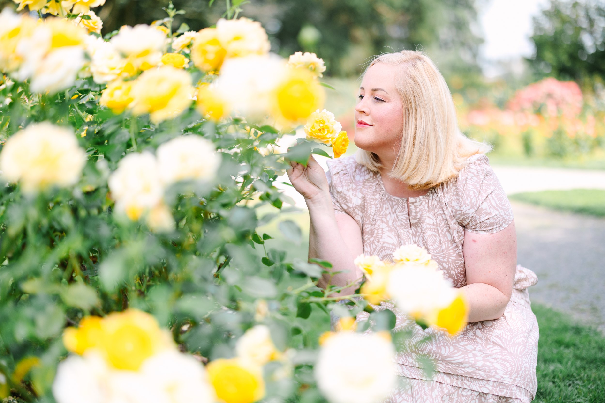 A woman in a pink floral dress smelling yellow roses in a garden during daytime.