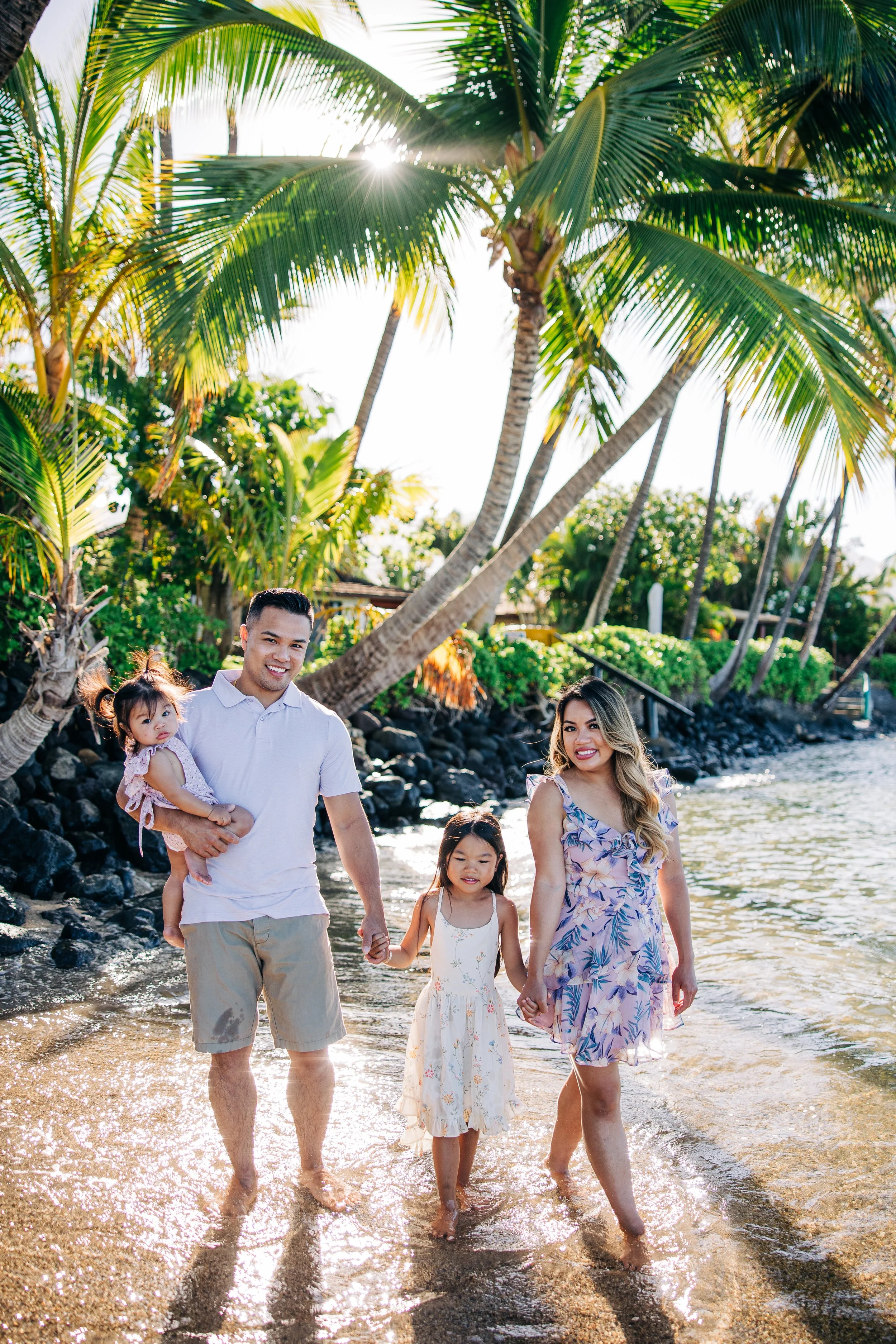 A happy multi-generational family walking hand in hand along the shoreline of a tropical beach during sunset, with palm trees and lush vegetation in the background.