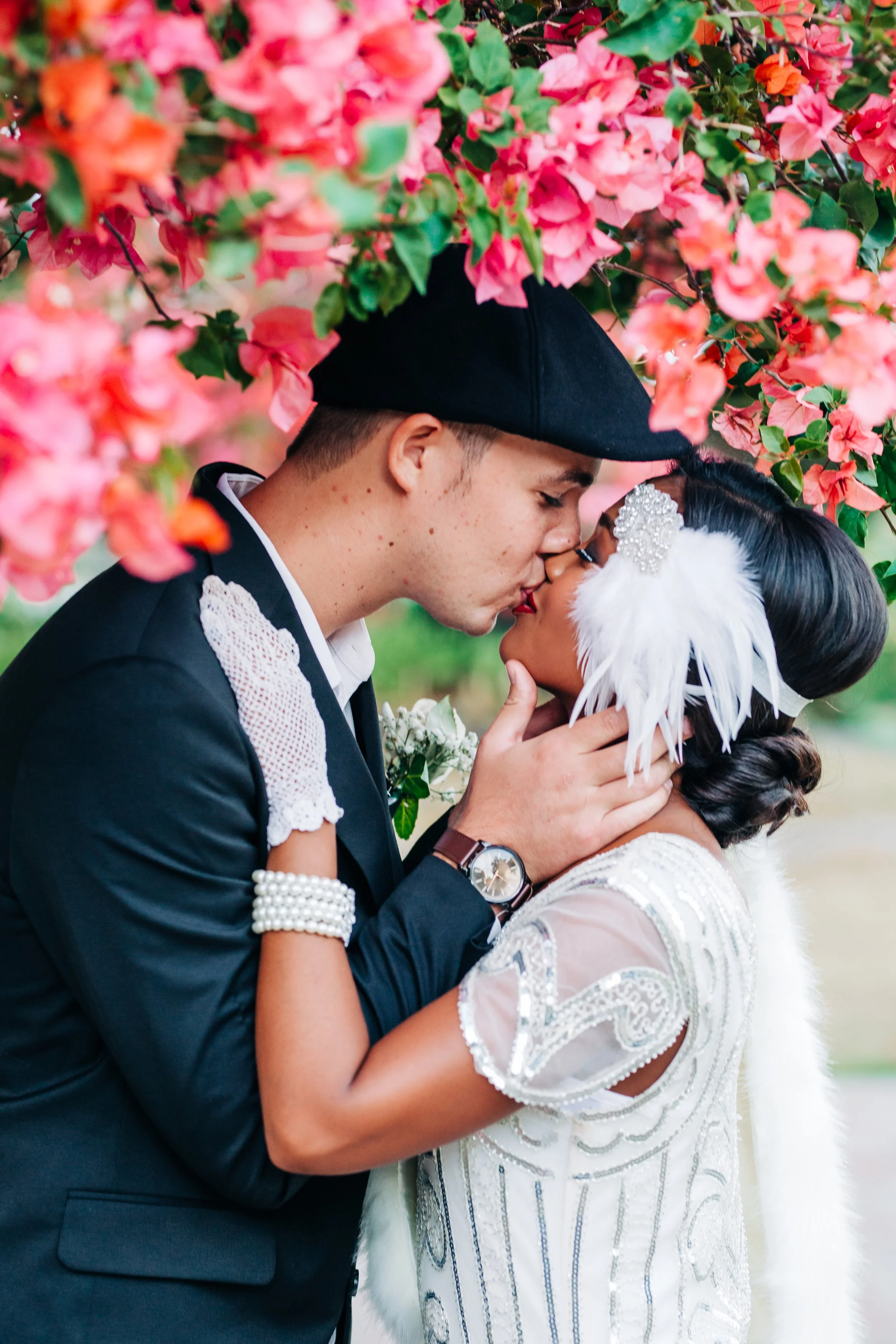 A wedding couple sharing a kiss under pink flowering bushes, with the groom wearing a black hat and a black suit, and the bride in a vintage style dress with feathers and pearl accessories.