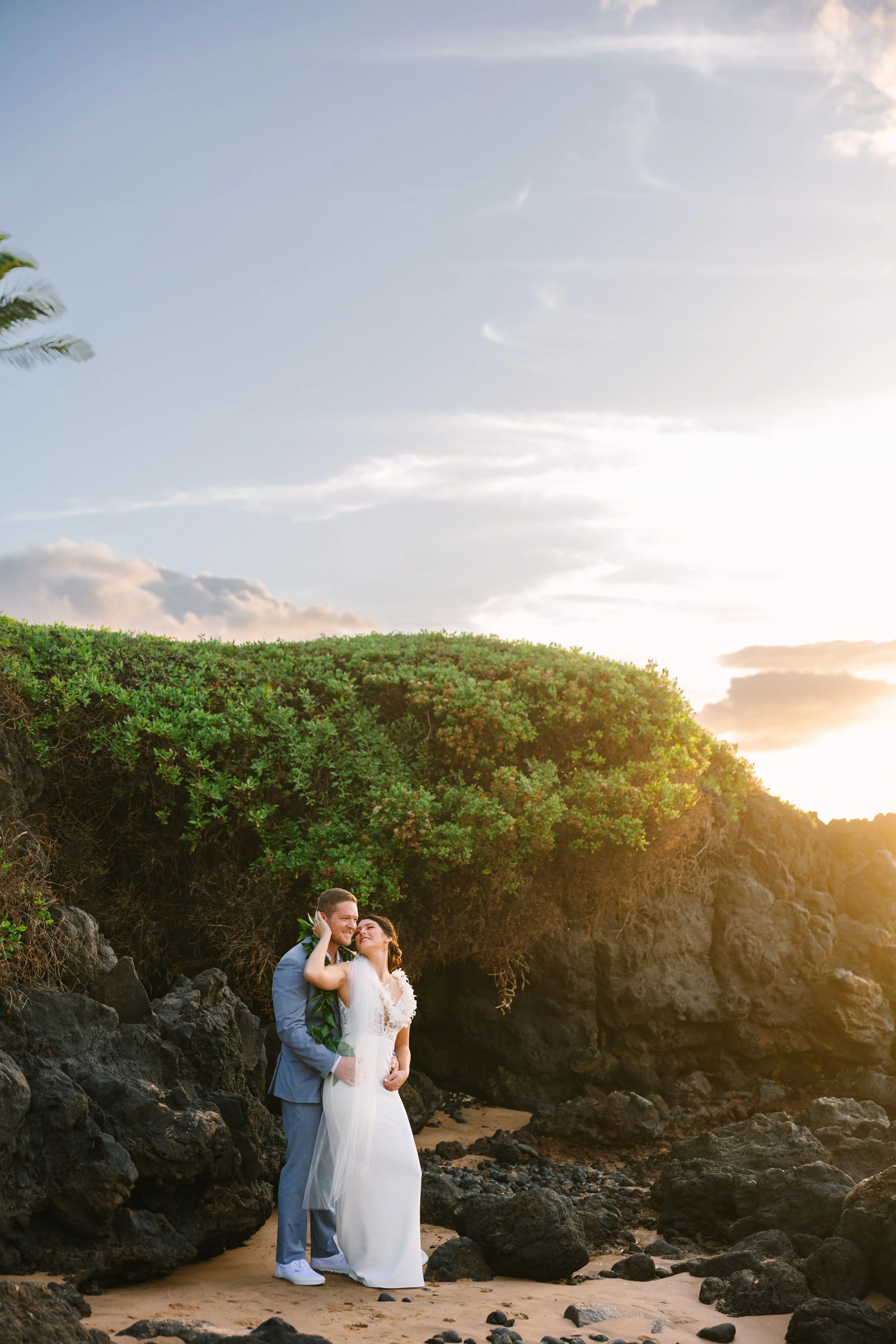 A couple in wedding attire standing on a rocky beach at sunset, embracing with a lush green hillside and sky in the background.