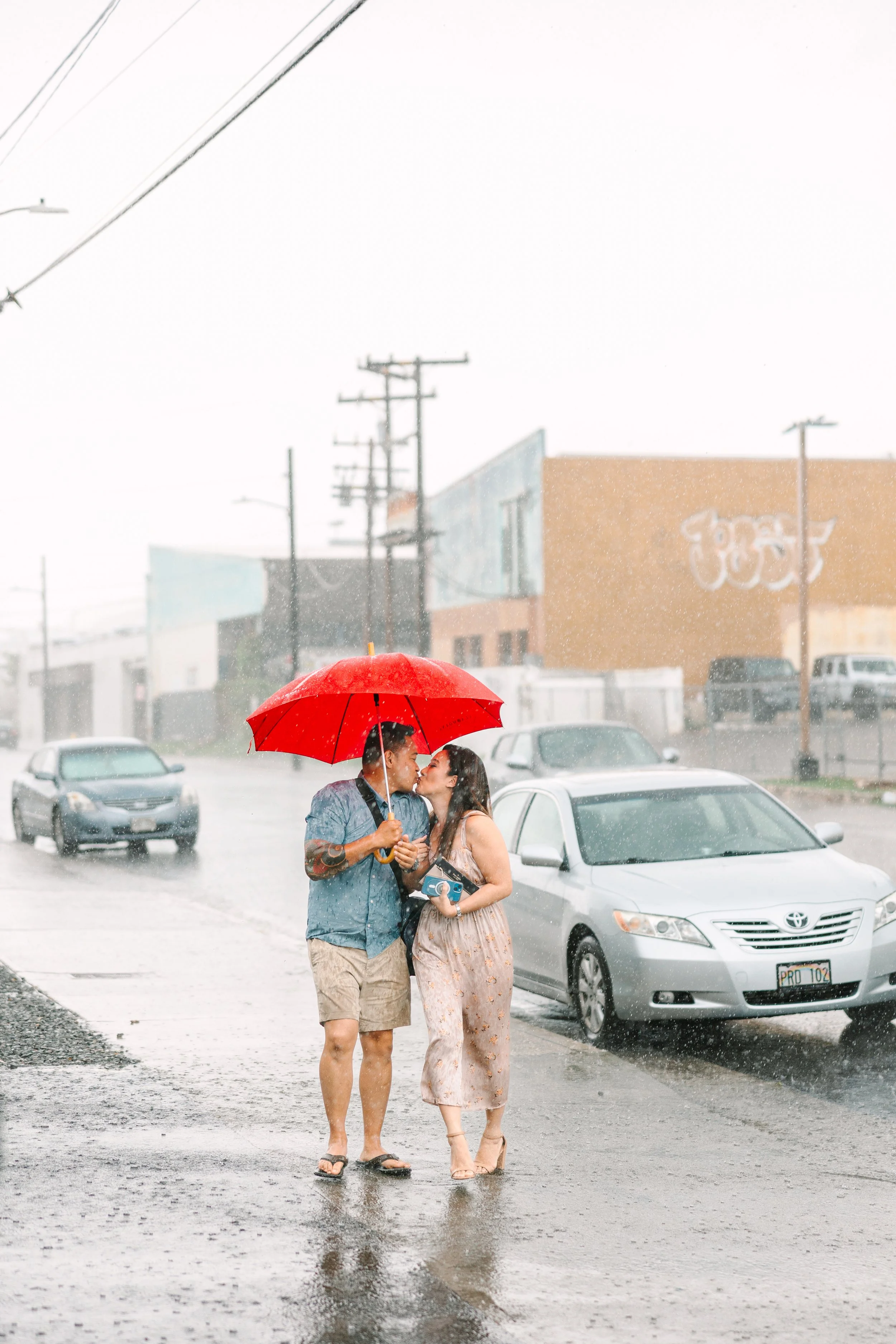 A couple stands close together in the rain under a red umbrella on a flooded street, kissing as rain pours down around them with cars parked and buildings in the background.