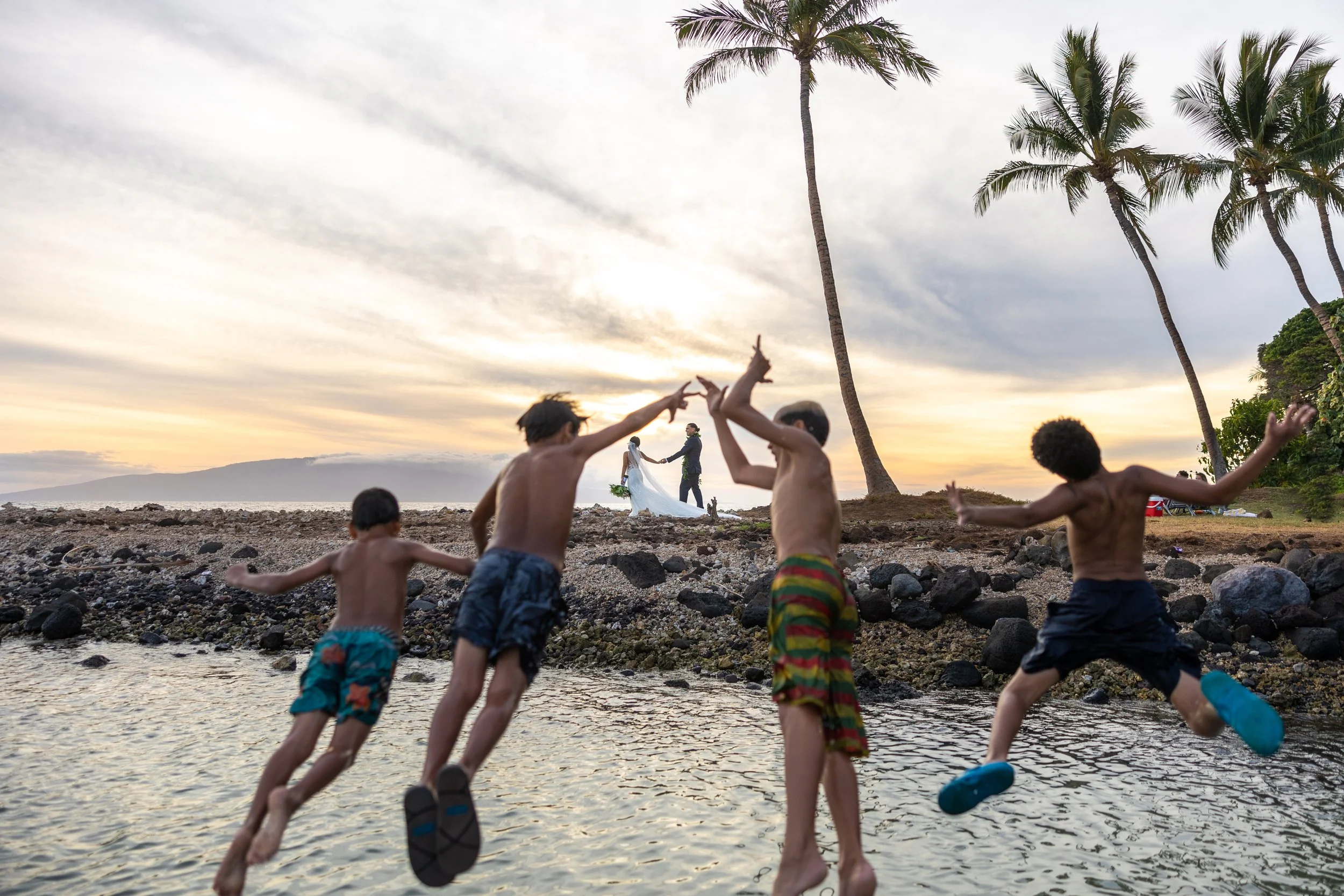 Children playing and running by the shore at sunset, with palm trees and a couple holding hands in the background.