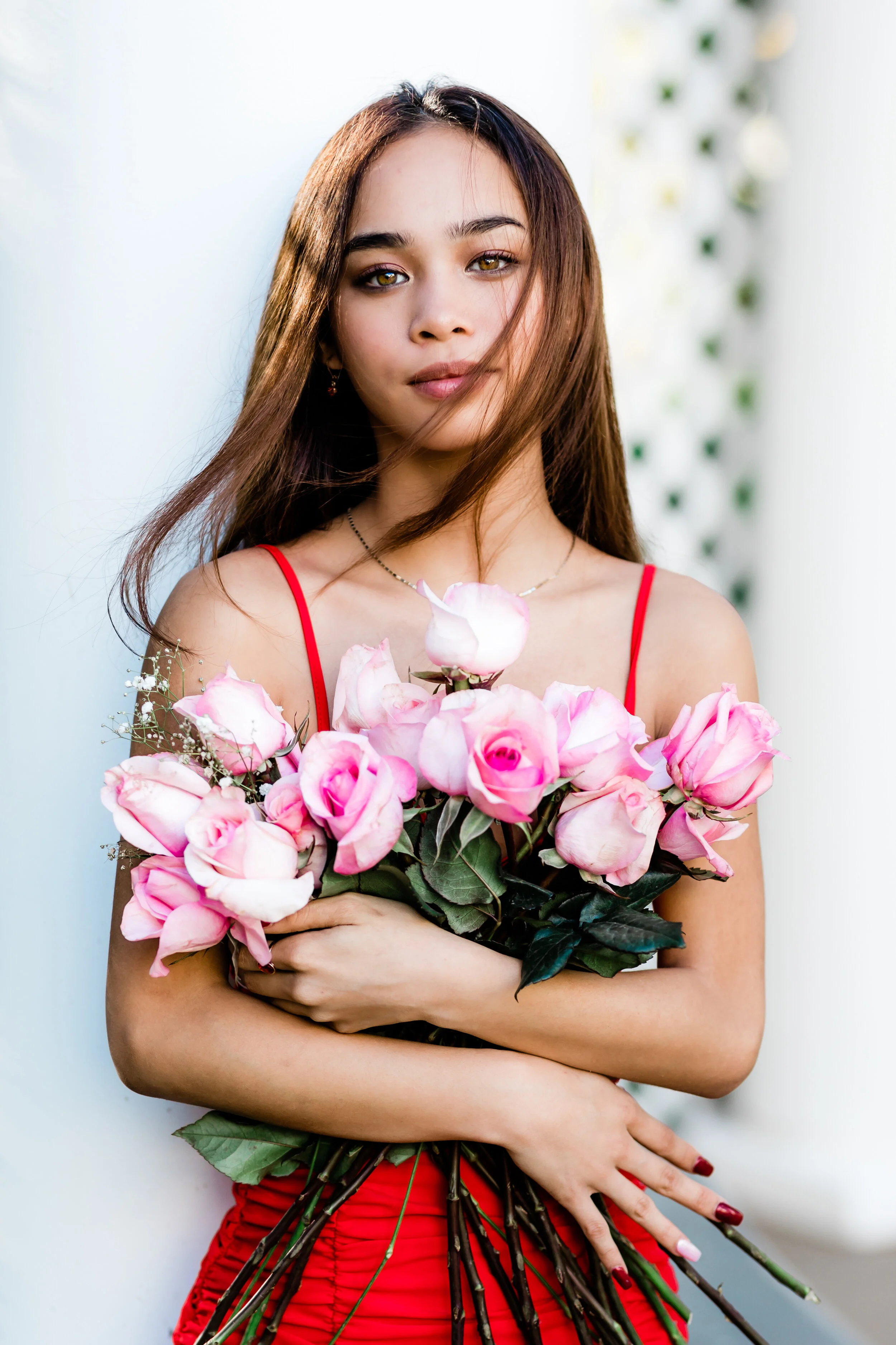 A woman with long brown hair and light brown eyes holding a large bouquet of pink roses and greenery, wearing a red dress, standing against a white background with some out-of-focus decorative elements.