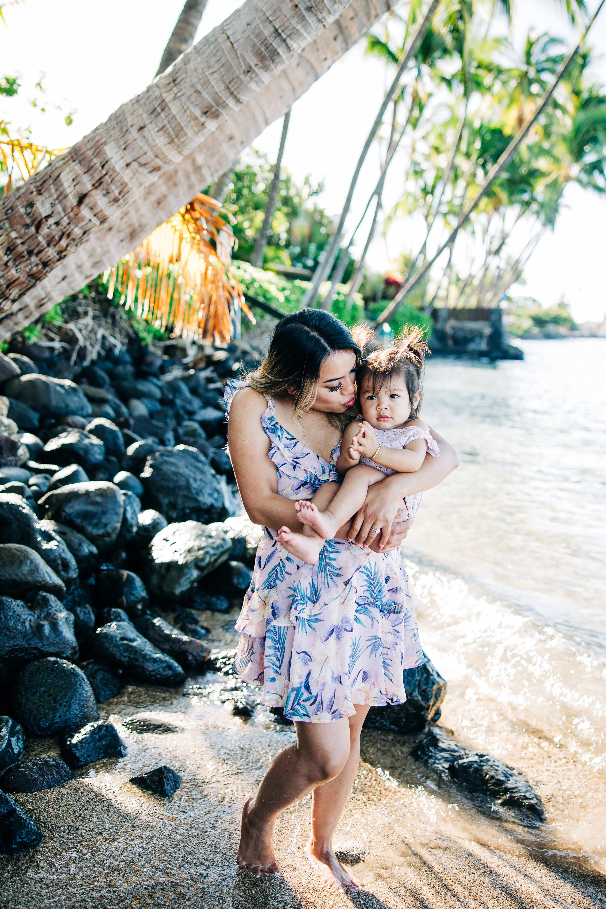 A woman holding a young girl while standing in shallow water near the rocky shoreline of a tropical beach with palm trees and a bright sky in the background.
