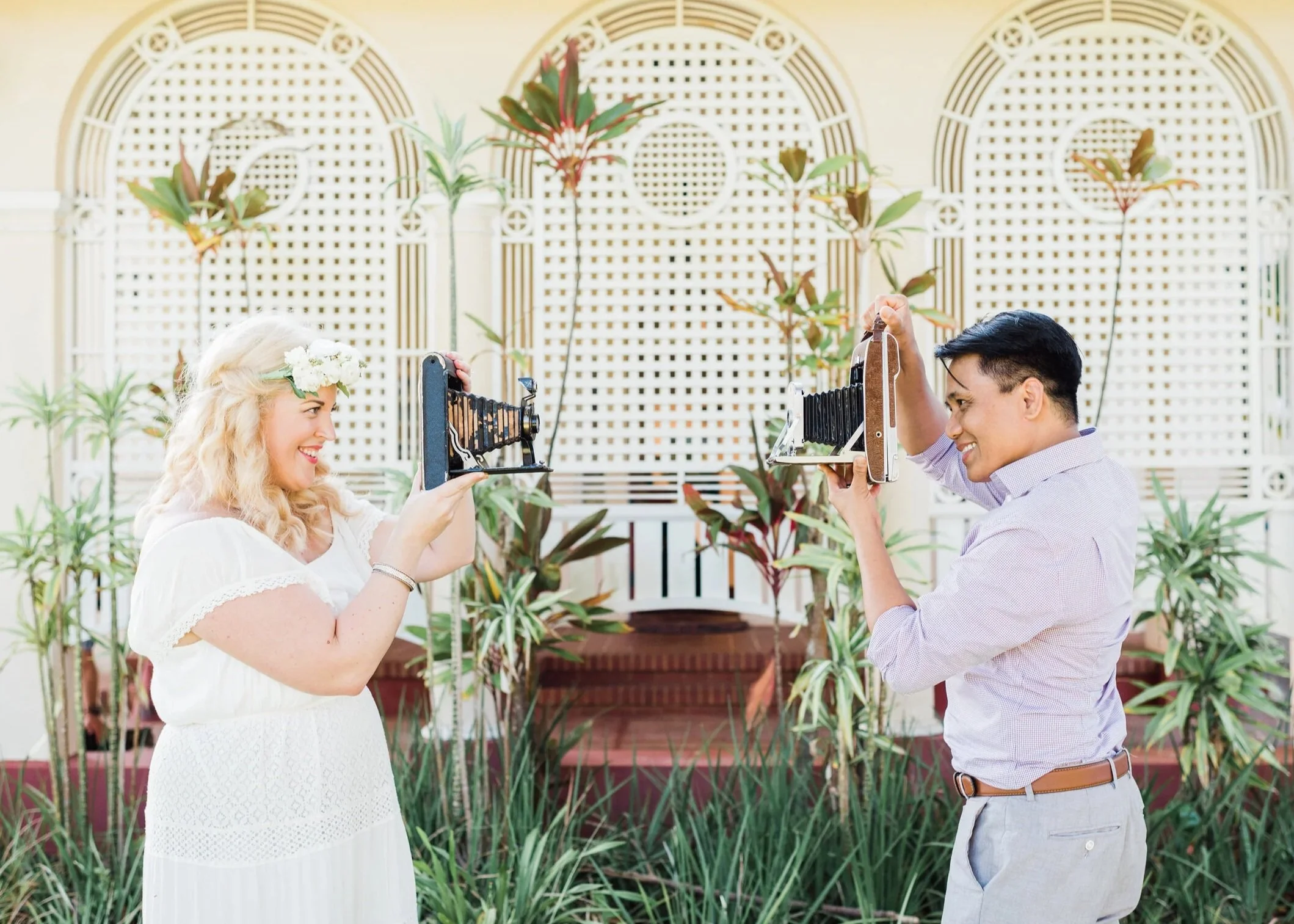 A woman with blonde curly hair and a flower crown holding a vintage camera, smiling at a man with short dark hair holding a similar vintage camera. They are outdoors in a garden with green plants and decorative white lattice panels in the background.