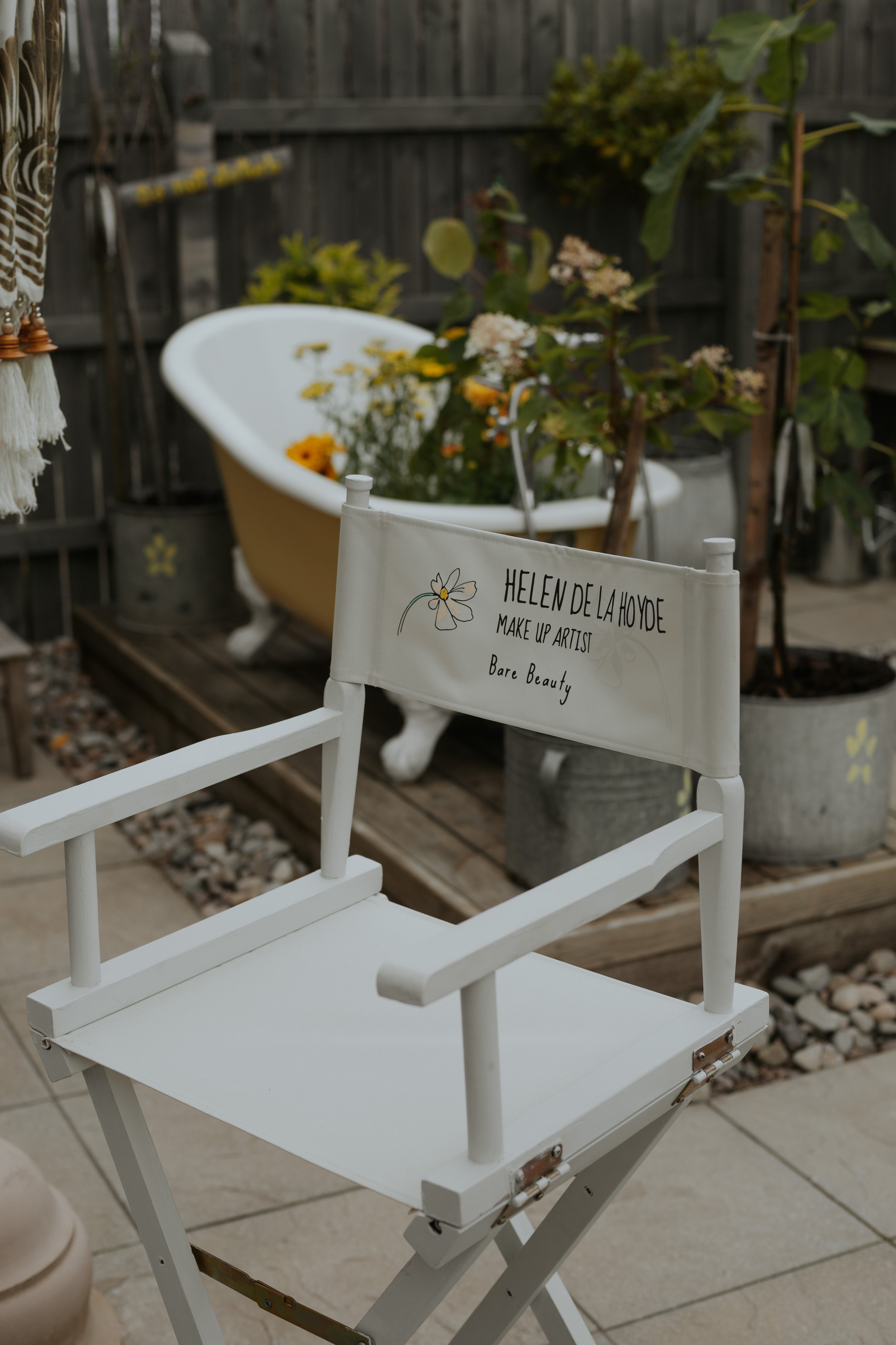 White outdoor chair with a sign that reads 'Helen de la Hoyde, Make Up Artist, Bare Beauty' in front of a vintage bathtub filled with flowers and plants, on a patio with a wooden fence in the background.