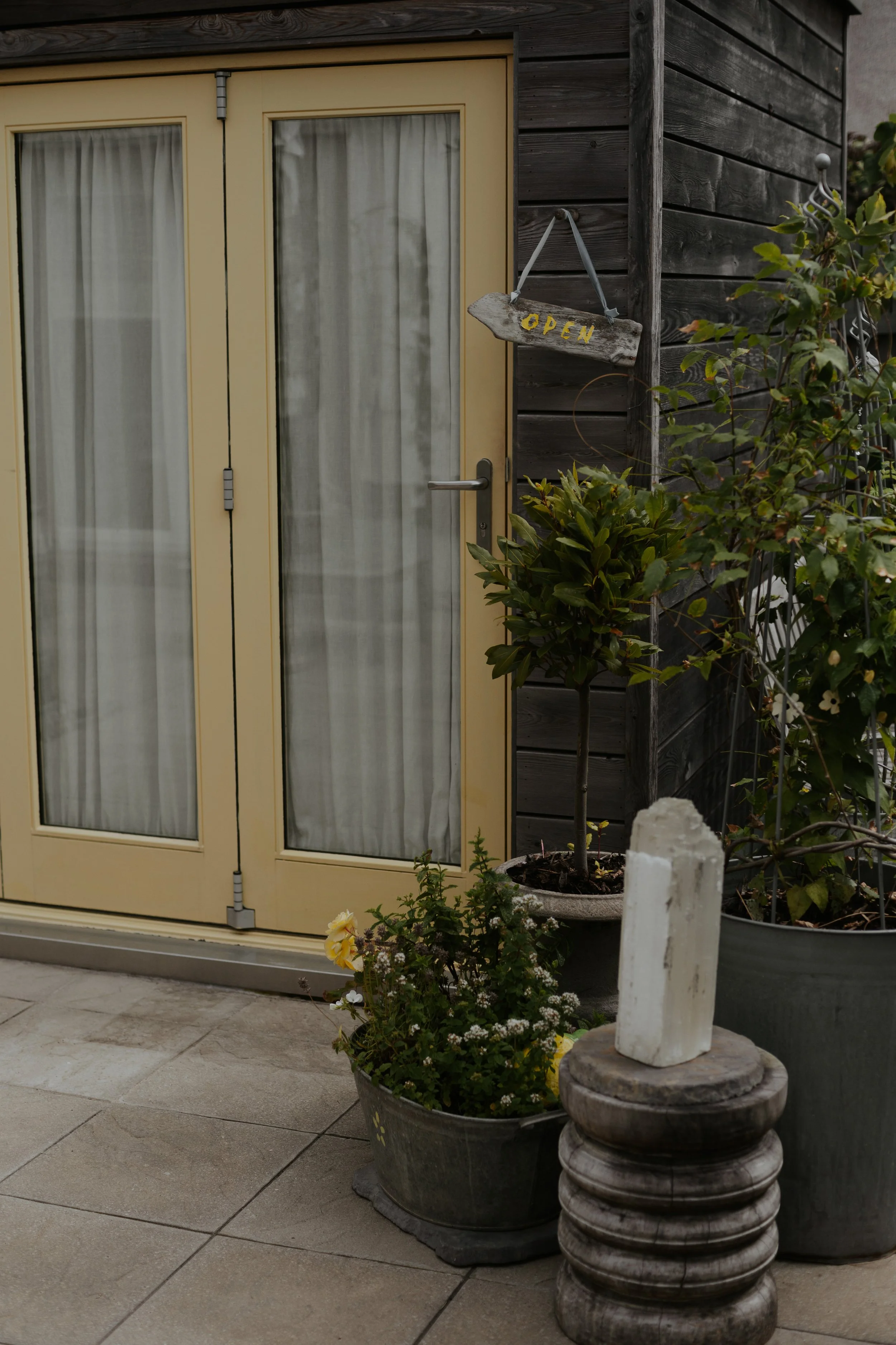 A yellow-framed glass door with white curtains, part of a building with dark wooden siding. Outside, there are potted plants, a small tree, and a weathered wooden 'Open' sign hanging on a corner of the wall, along with decorative stone planters and a white fence post.