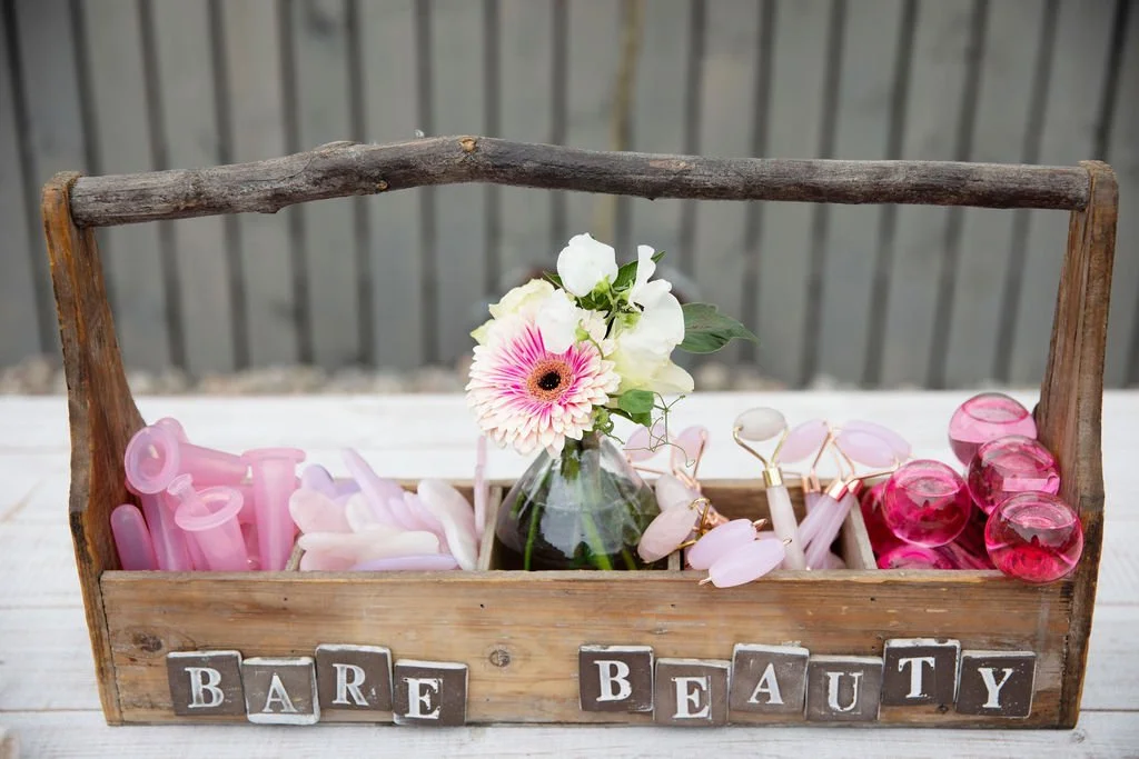 Wooden tray with pink and white beauty tools and a small flower vase with white and pink flowers, spelling out "Bare Beauty".