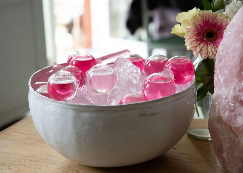 A white bowl filled with ice cubes and pink gel-like spheres, placed on a wooden surface with a flower arrangement nearby.
