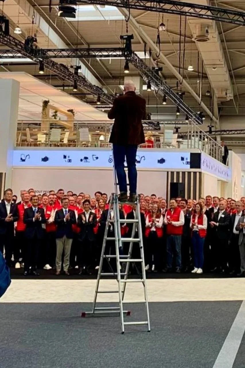 Ralf Milde fotografiert von einer Leiter aus eine große Firmengruppe in einer Messehalle