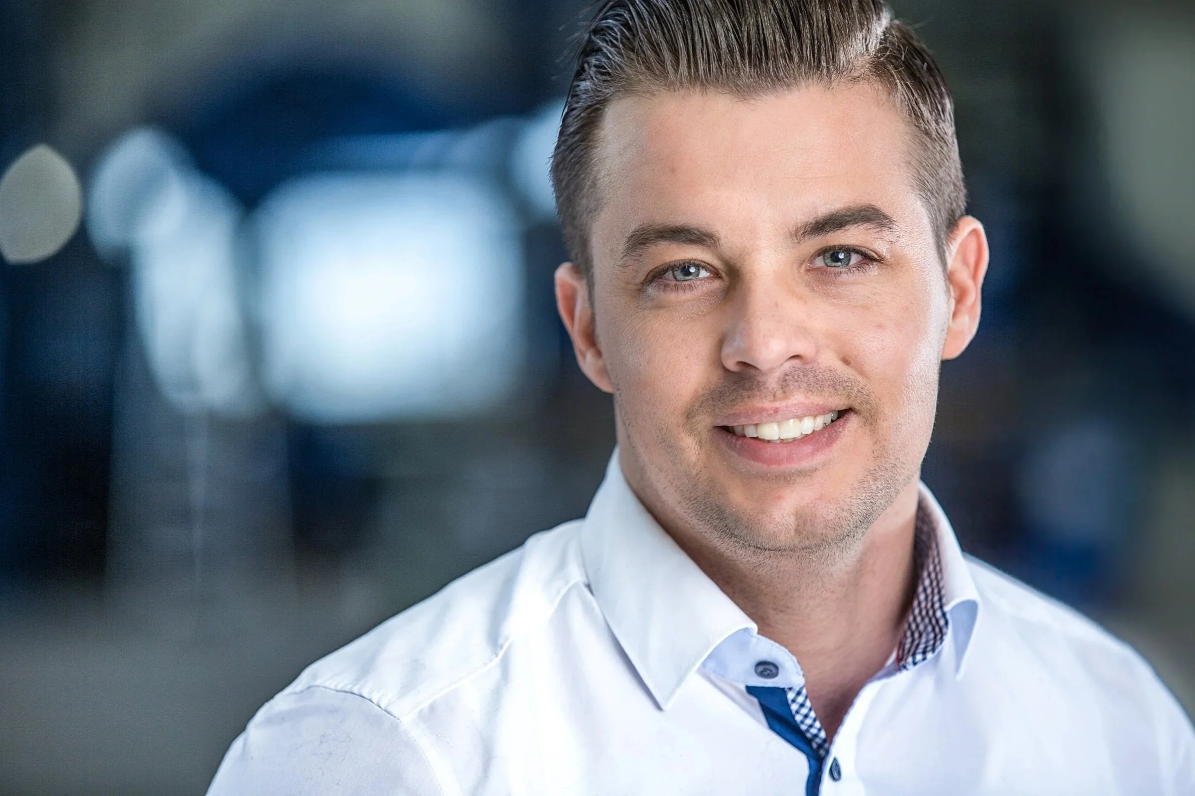 Businessportrait eines Mitarbeiters mit dunklen Haaren und weißem Hemd, professioneller Cinematic-Look mit unscharfem Industrie-Hintergrund