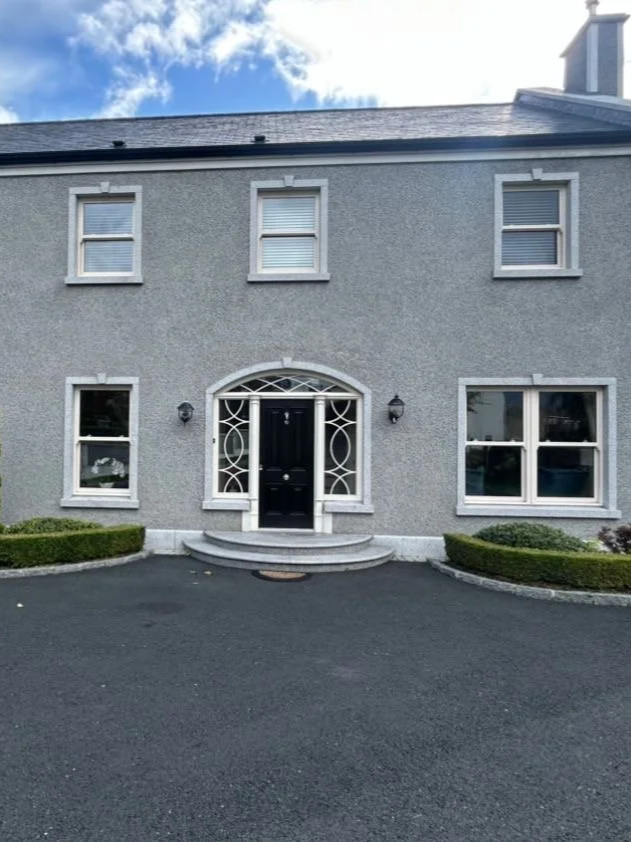Front exterior of a two-story grey house with white window frames, a black front door with decorative glass, surrounded by green shrubs and a paved driveway.