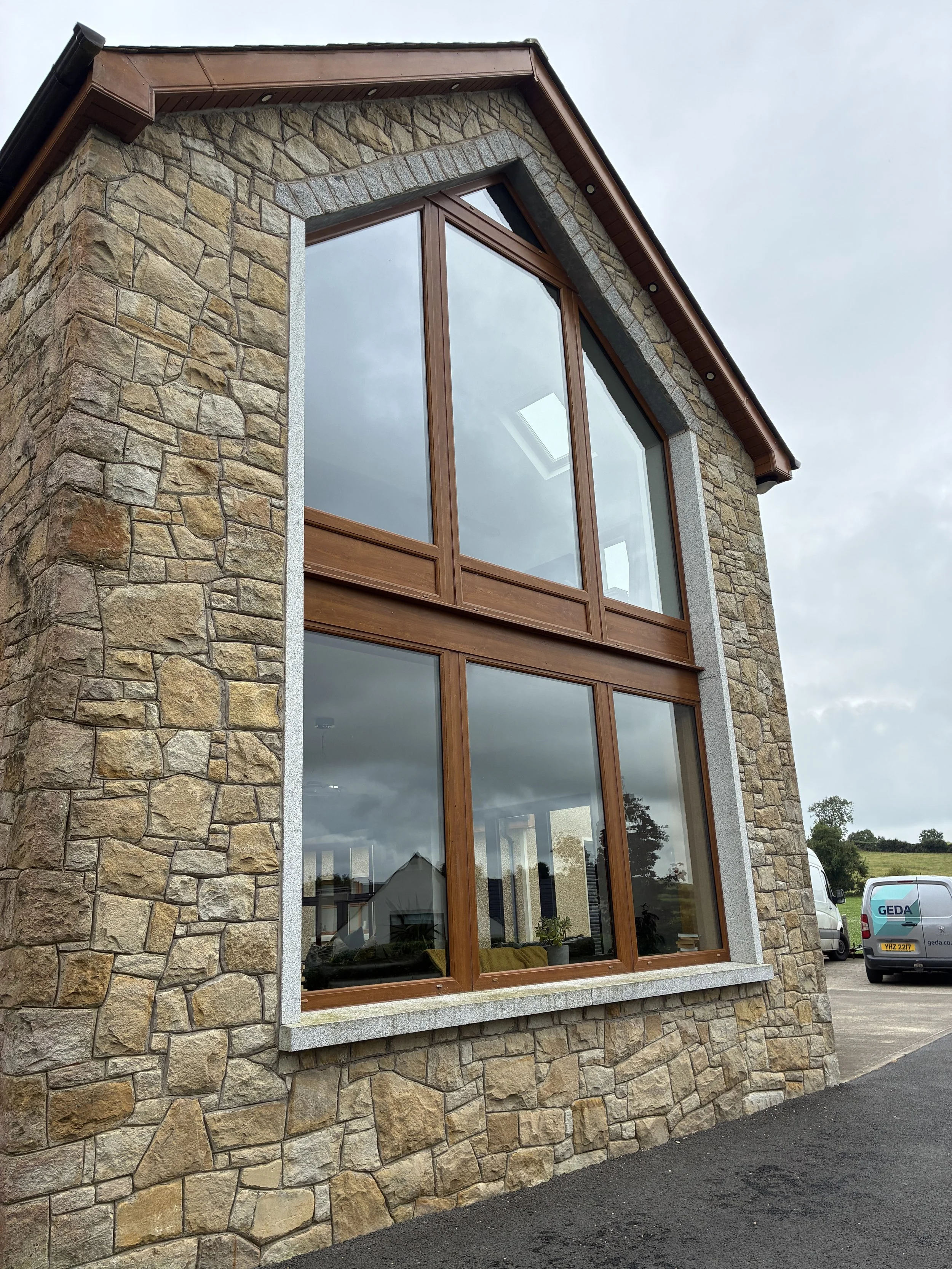 A stone house with large brown-framed windows on a cloudy day, with a view of the interior and parked vehicles outside.