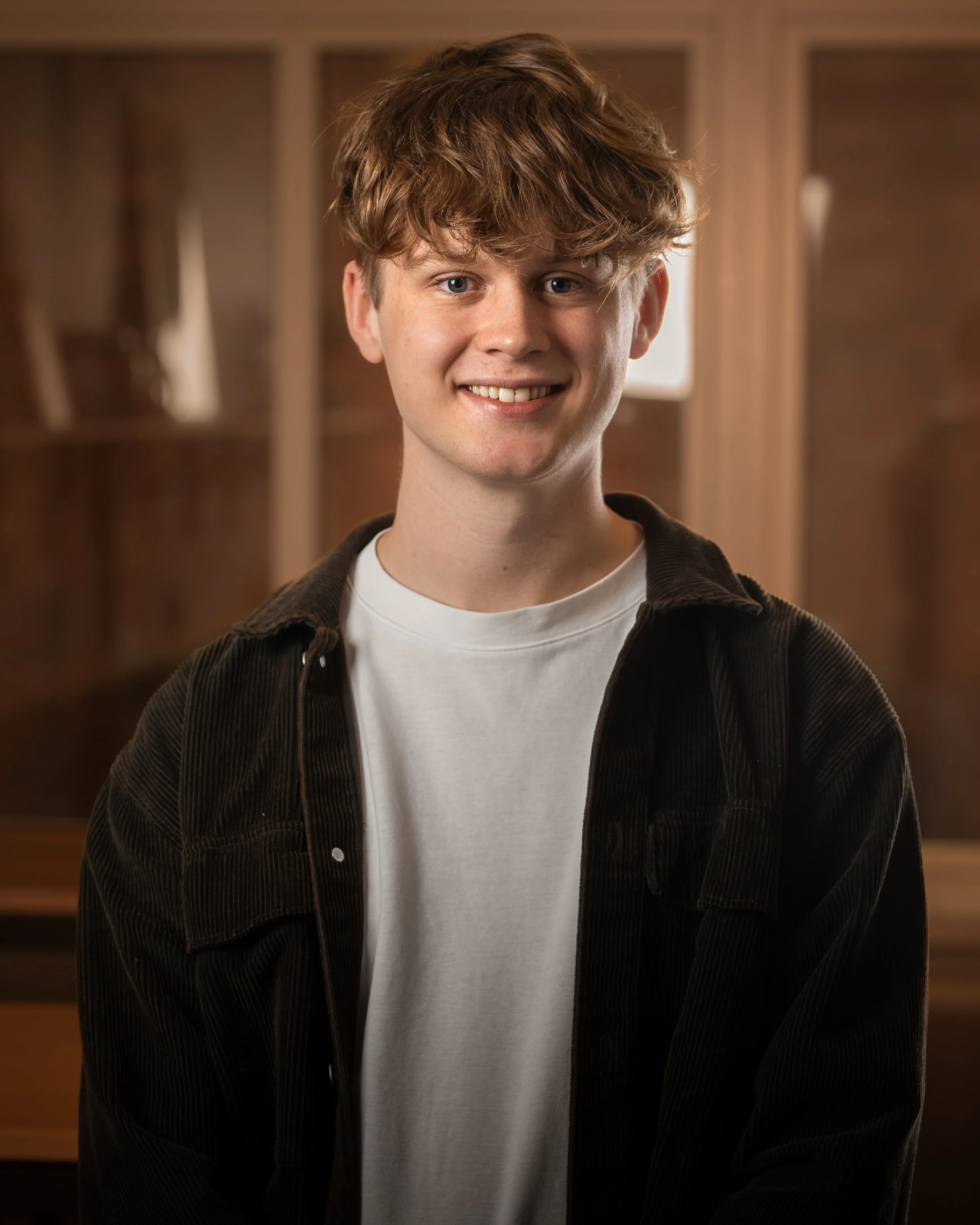 Ung man med brunt lockigt hår, vita tänder, svart jacka och vit t-shirt, ler i en inomhusmiljö.