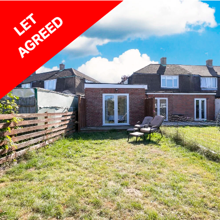 Backyard with a small red brick building, outdoor chairs, a wooden fence, and neighboring houses in the background under a partly cloudy sky.