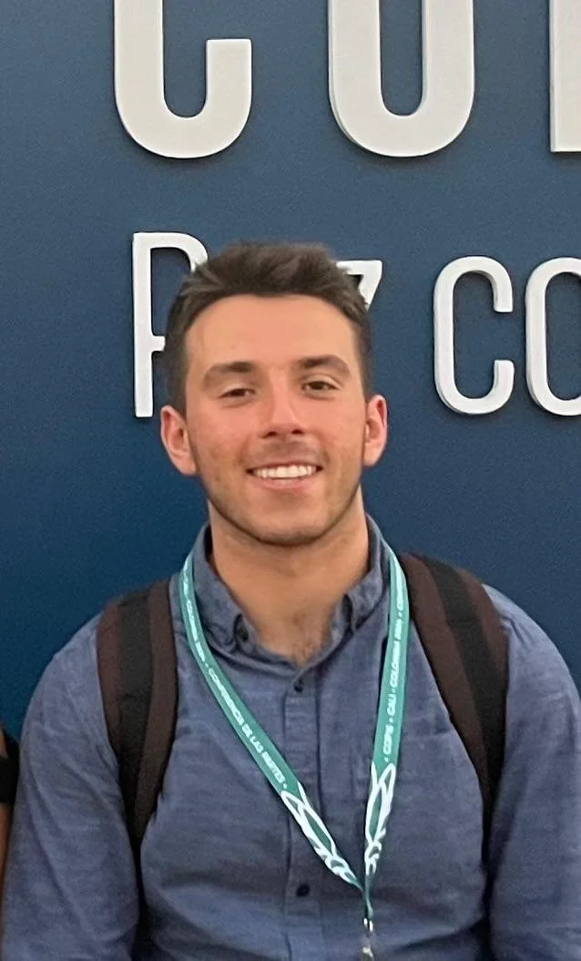 A young man smiling in front of a blue wall with white large letters, wearing a blue shirt, a backpack, and a teal lanyard around his neck.