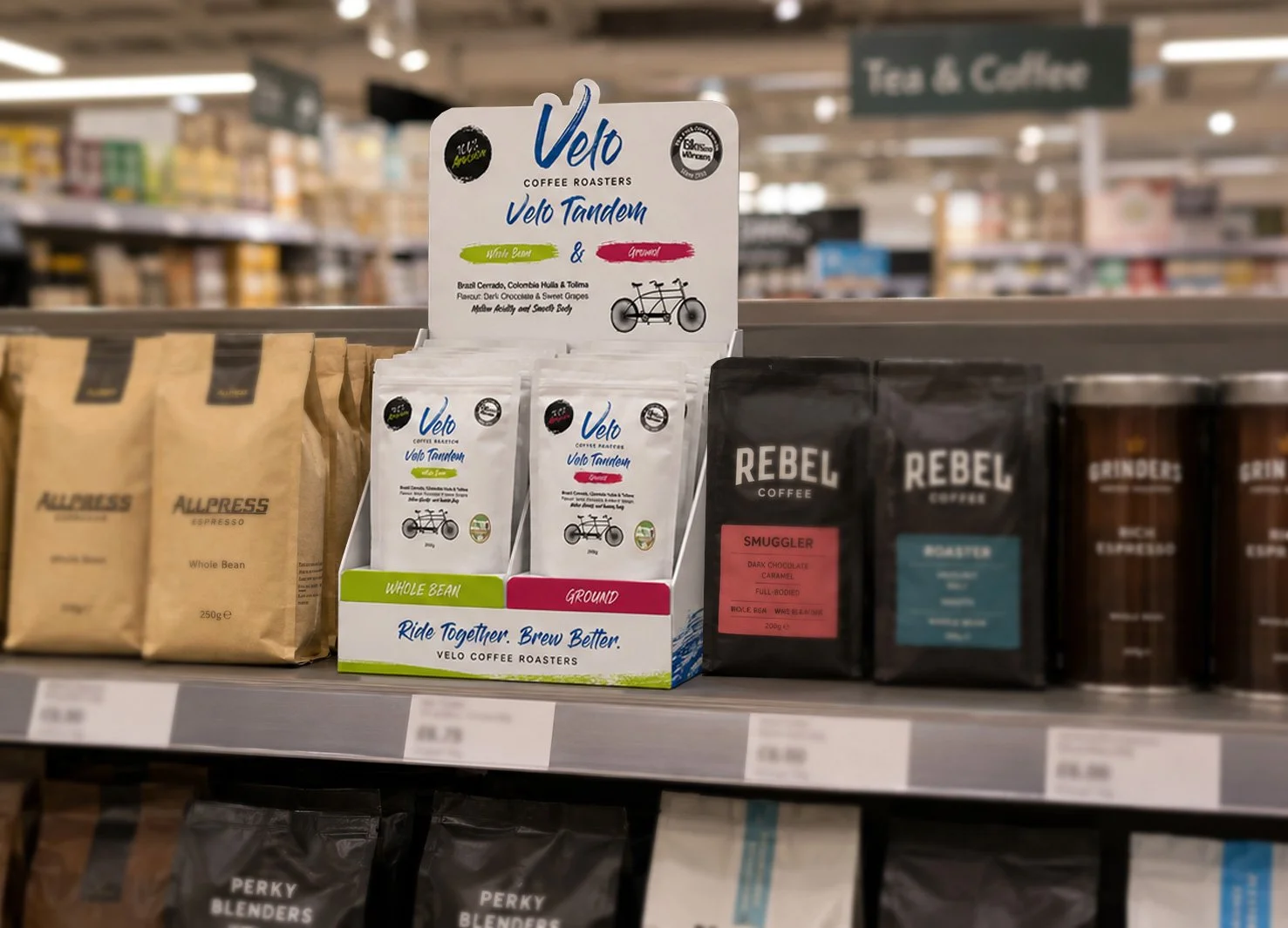 Display of coffee bags in a grocery store. The focus is on Velo Tandem Coffee Roasters' whole bean and ground coffee packages, with Rebel Coffee and Brinders coffee bags visible in the background.