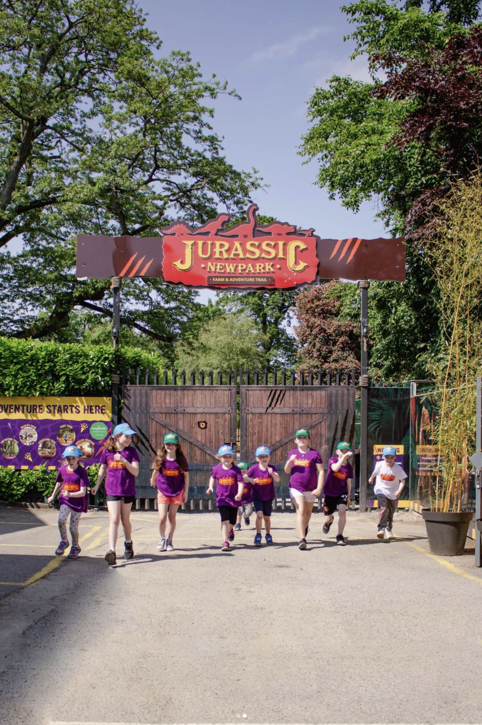 Children walking out of Jurassic New Park entrance, a theme park, with a sign that reads 'Jurassic New Park' and a logo of dinosaurs. The children are wearing purple shirts and blue caps, and there are trees and a gate in the background.