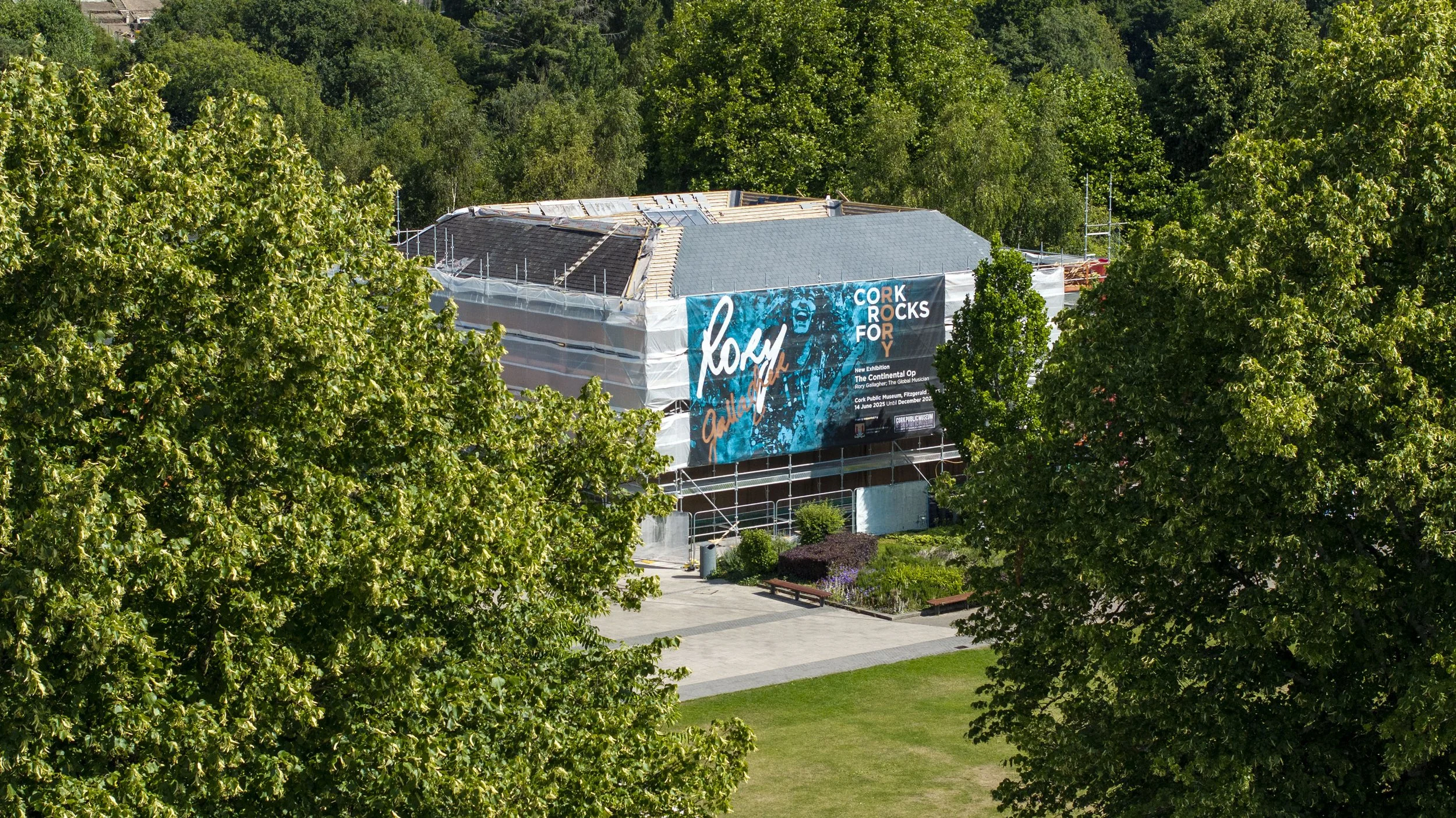 Construction scaffolding around a building with a large Cork Rocks for Rory poster, surrounded by green trees and a park area.