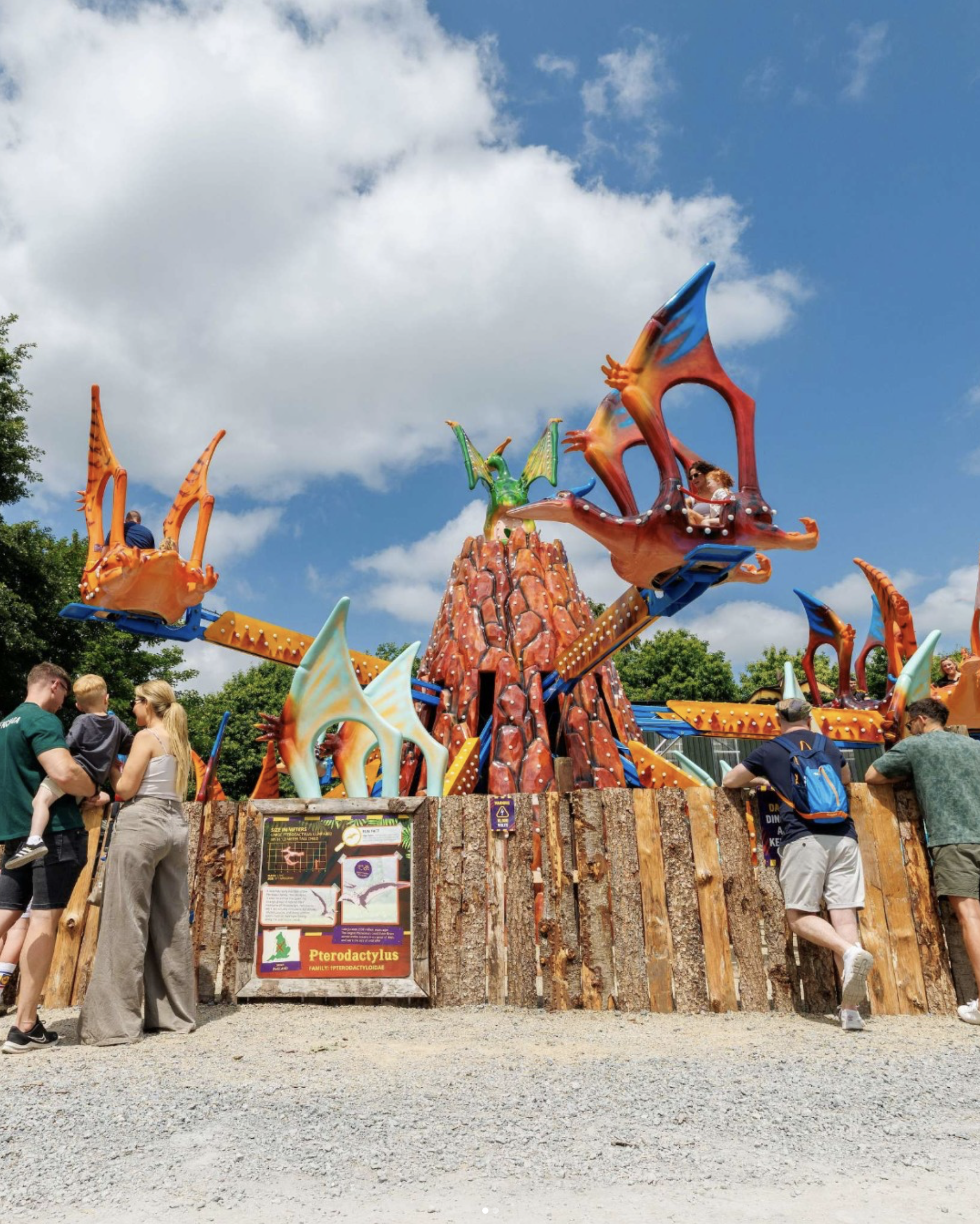 Colorful amusement park ride featuring dragon-themed swings and a volcano-style central structure, with people waiting and riding on a sunny day.
