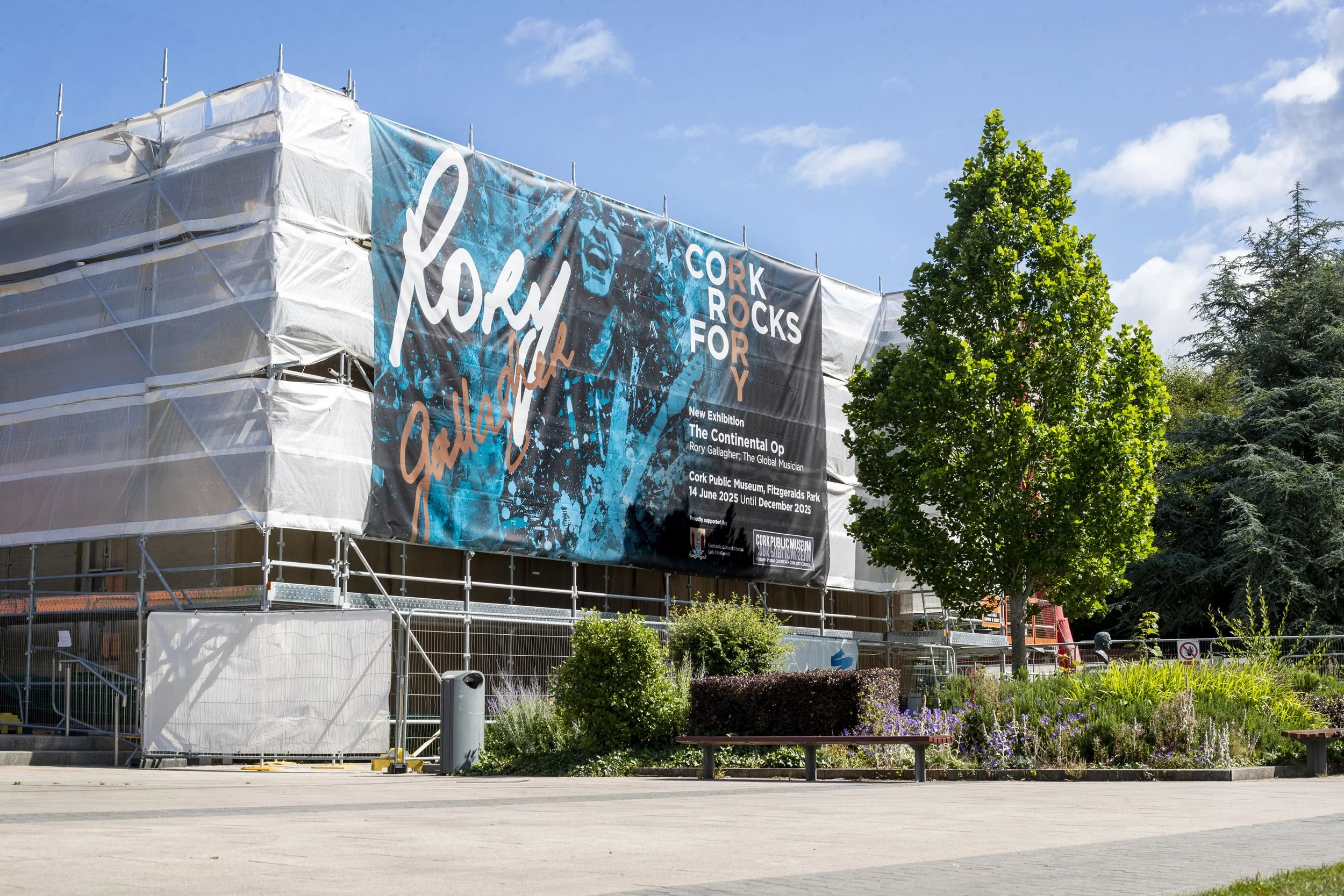 Construction scaffolding covered with a large banner for an exhibition titled Cork Rocks for Rory Gallagher, at Cork Public Museum, Fitzgerald's Park, with trees and benches in the foreground.