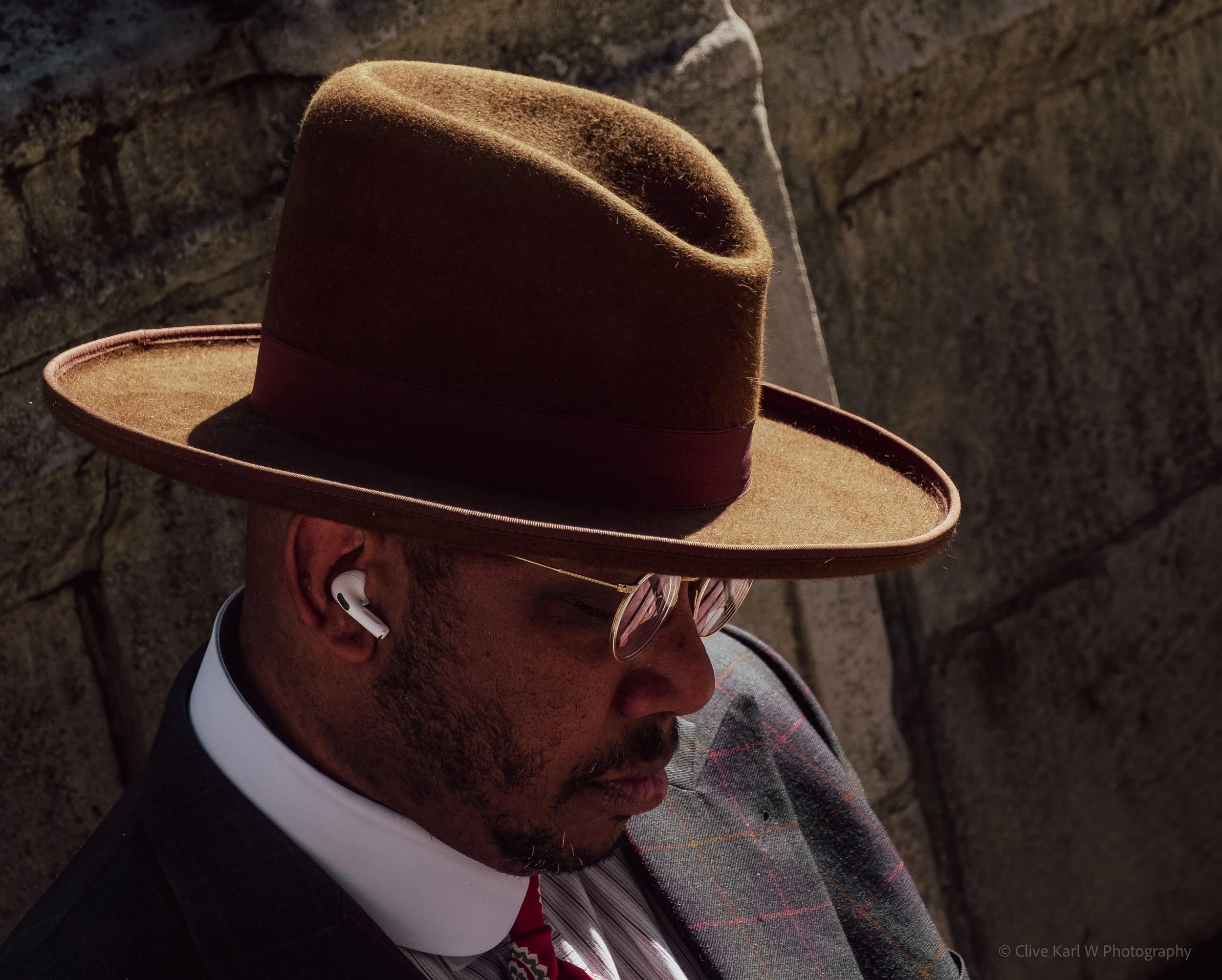 Close-up of a man wearing a brown felt cowboy hat, glasses, and a plaid shirt, outdoors near a stone wall.