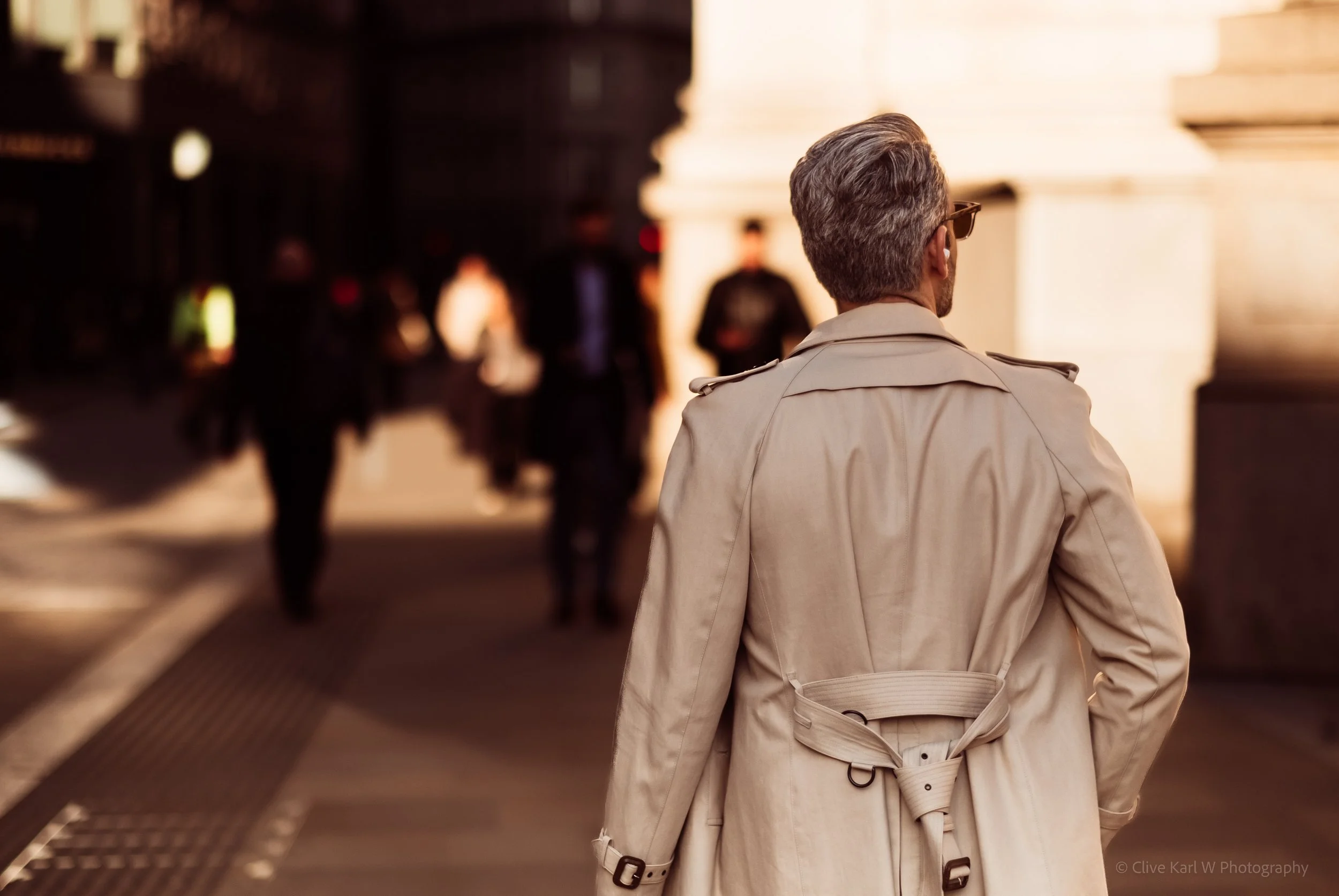 A man wearing a beige trench coat, seen from behind, walking outdoors in a city at dusk.