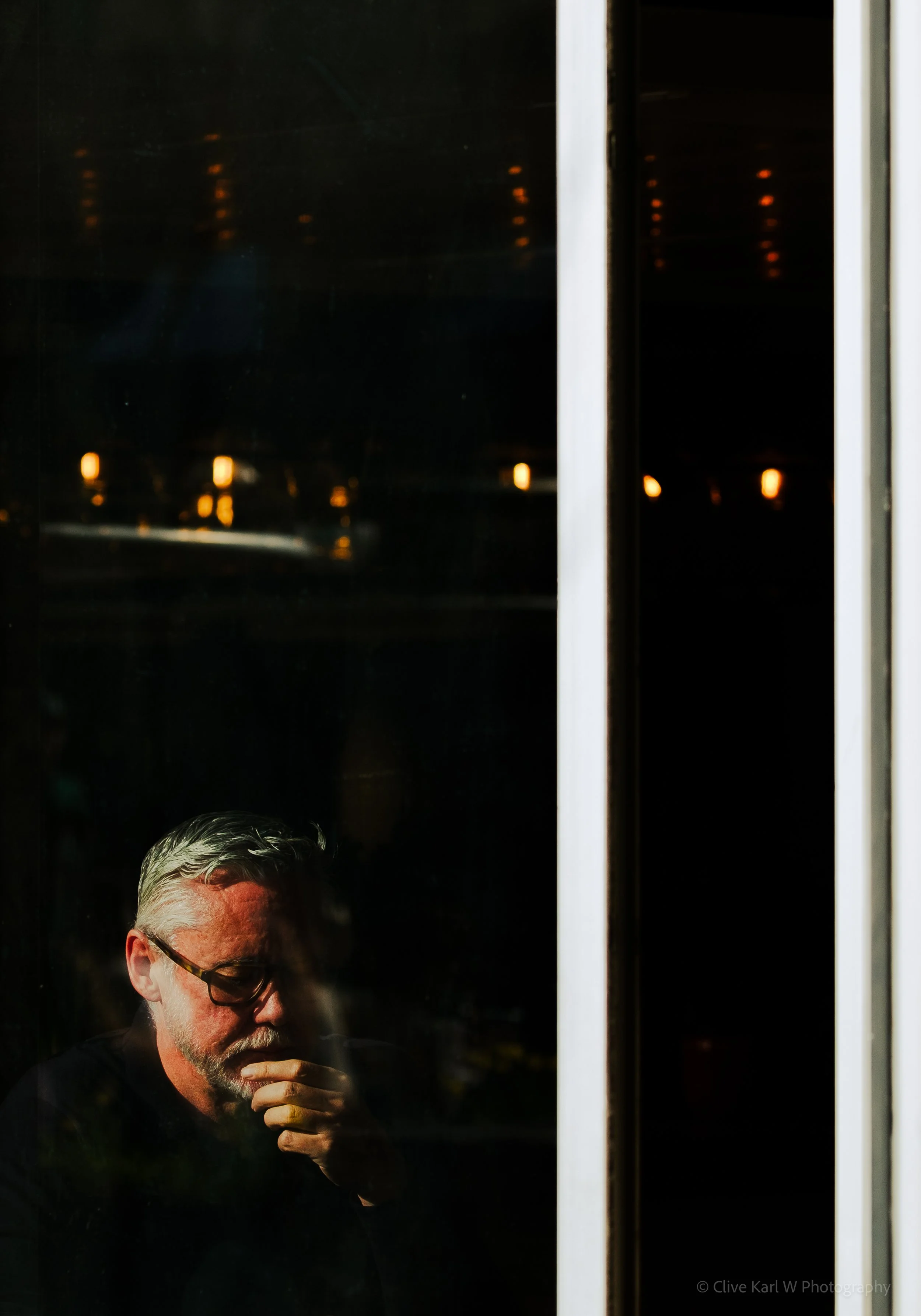 Reflection of a man with gray hair, beard, glasses, wearing a dark shirt, taking a sip from a cup, seen through a glass window at night with dark background and orange lights.