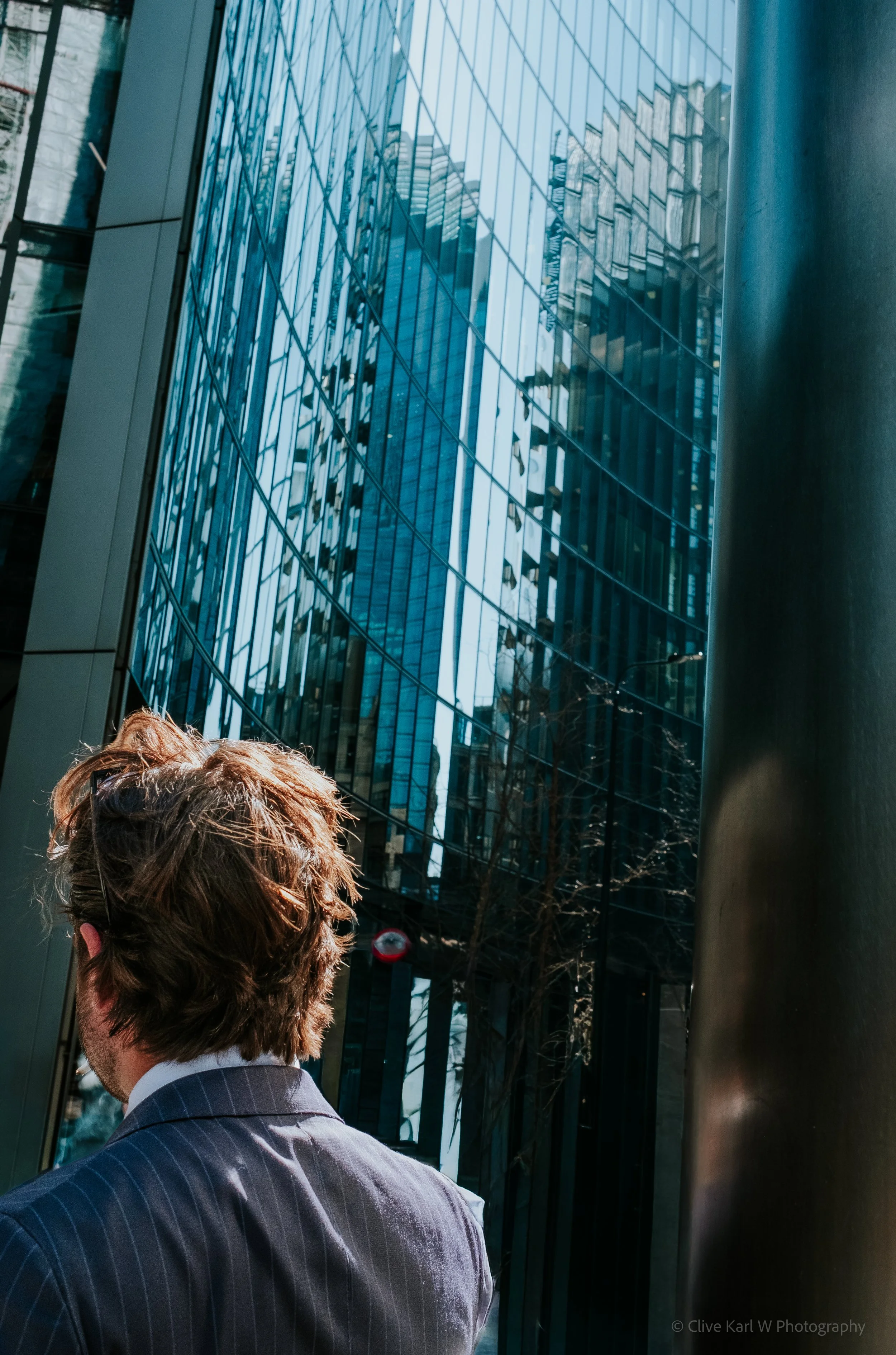 A person with curly brown hair wearing a suit in front of reflective glass windows of a modern high-rise building.