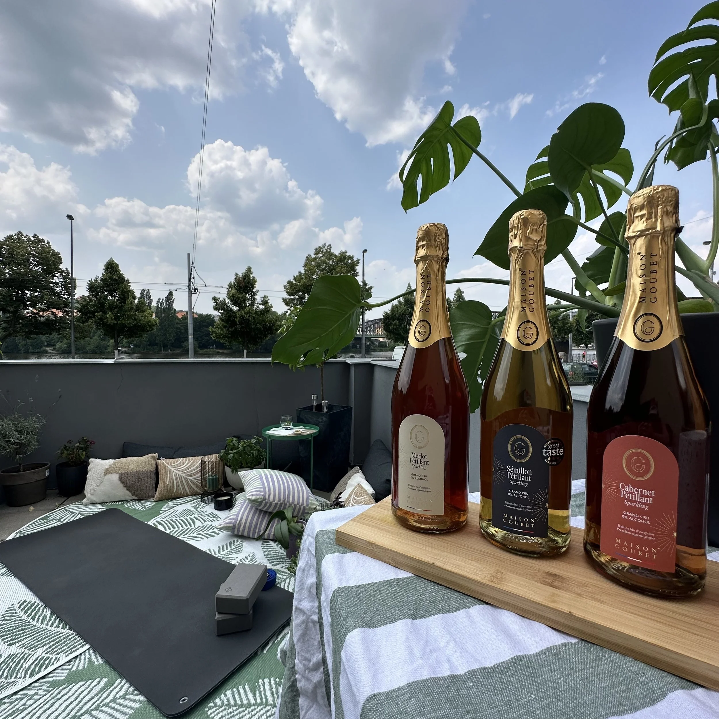 An outdoor balcony with a table covered by a leafy patterned cloth, decorated with pillows, plants, and a laptop. In the foreground, a wooden tray holds three bottles of sparkling wine with gold foil caps and labels, placed beside a potted climbing plant.