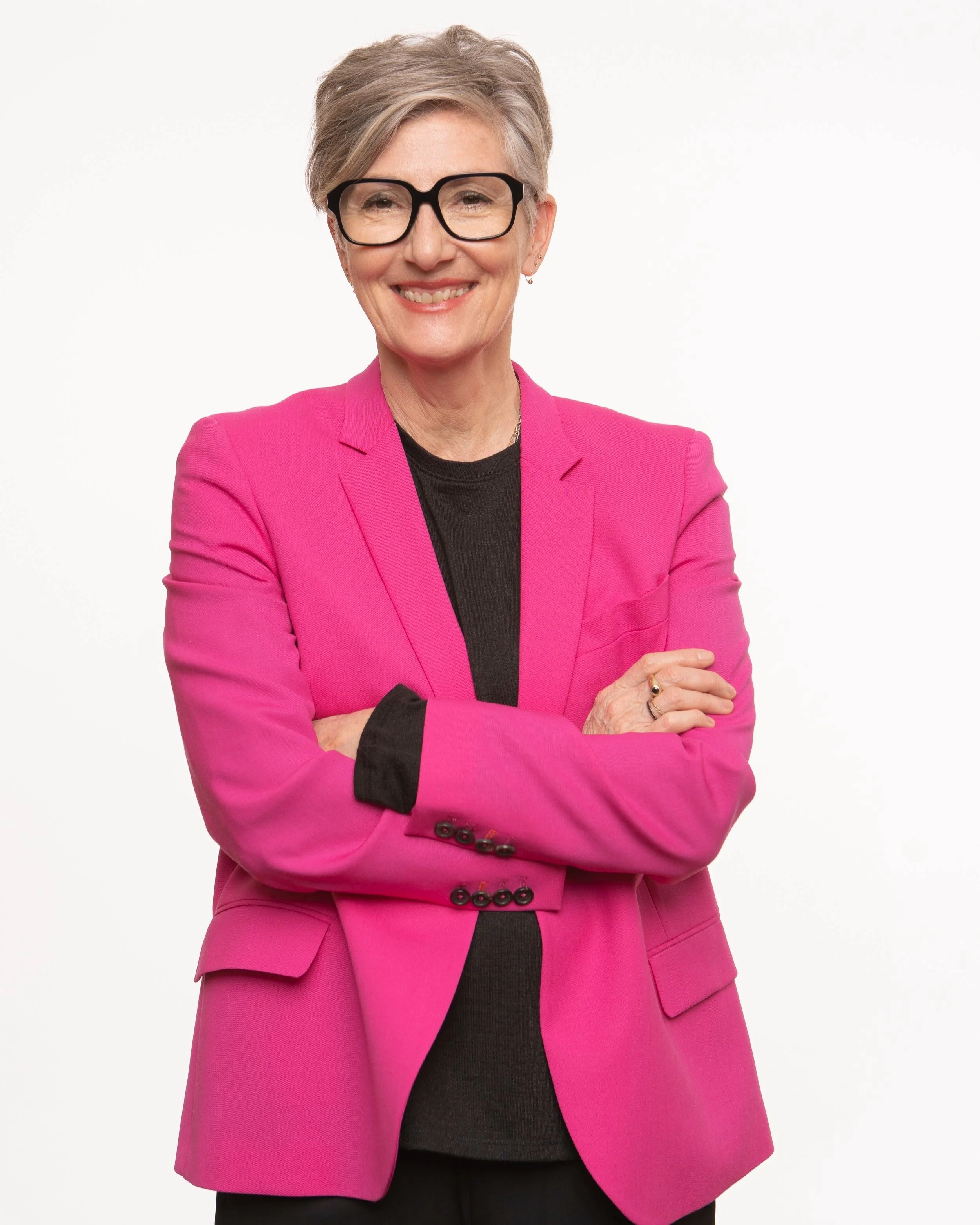 Jeanette Bronée with short gray hair, black glasses, and earrings, smiling, dressed in a bright pink blazer over a black top, standing with arms crossed against a plain white background.