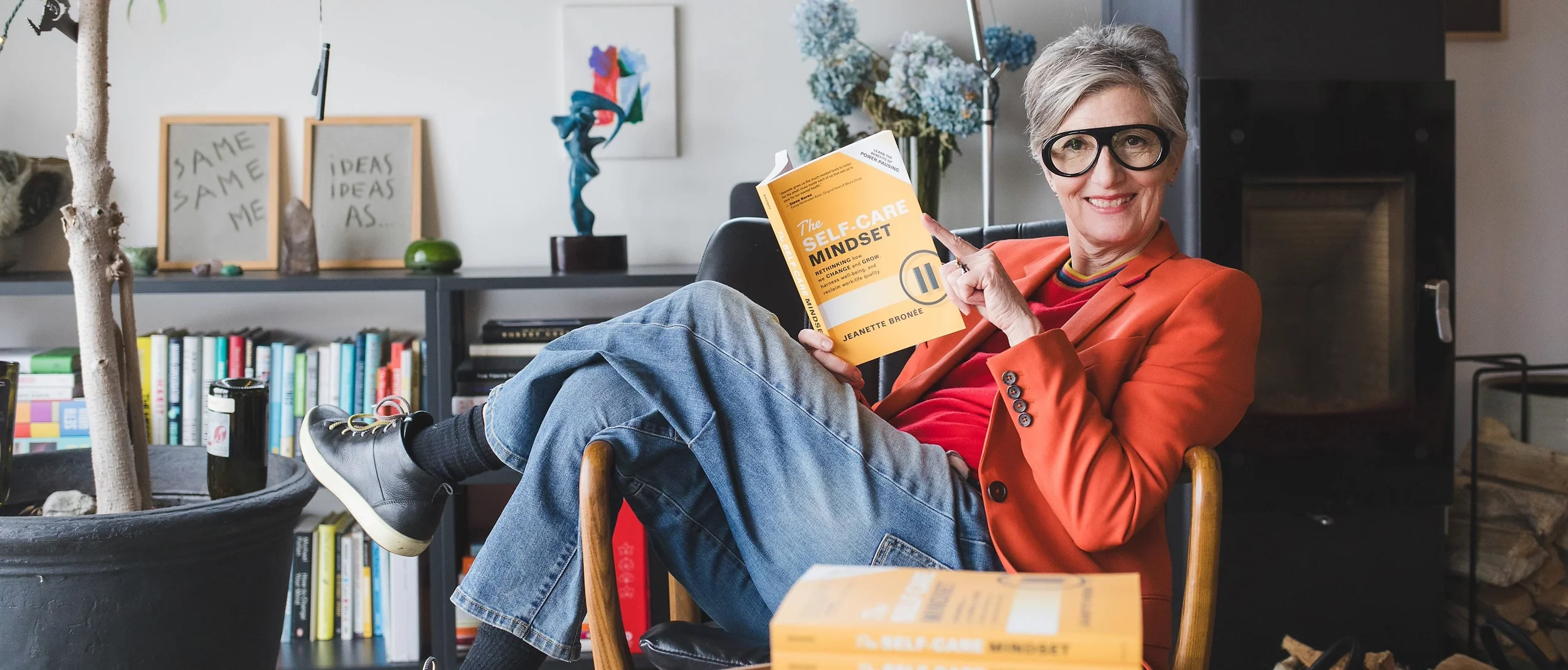 An older woman with short gray hair and glasses, wearing a red shirt and orange blazer, sitting in a chair, smiling, and holding a book titled "The Self-Care Mindset" in a cozy, well-decorated room with bookshelves, artwork, and plants.